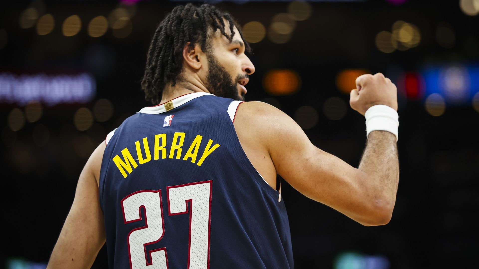 A basketball player wearing a navy blue jersey with yellow "MURRAY" and number 27, raising a fist in celebration during a game with blurred stadium lights in the background.