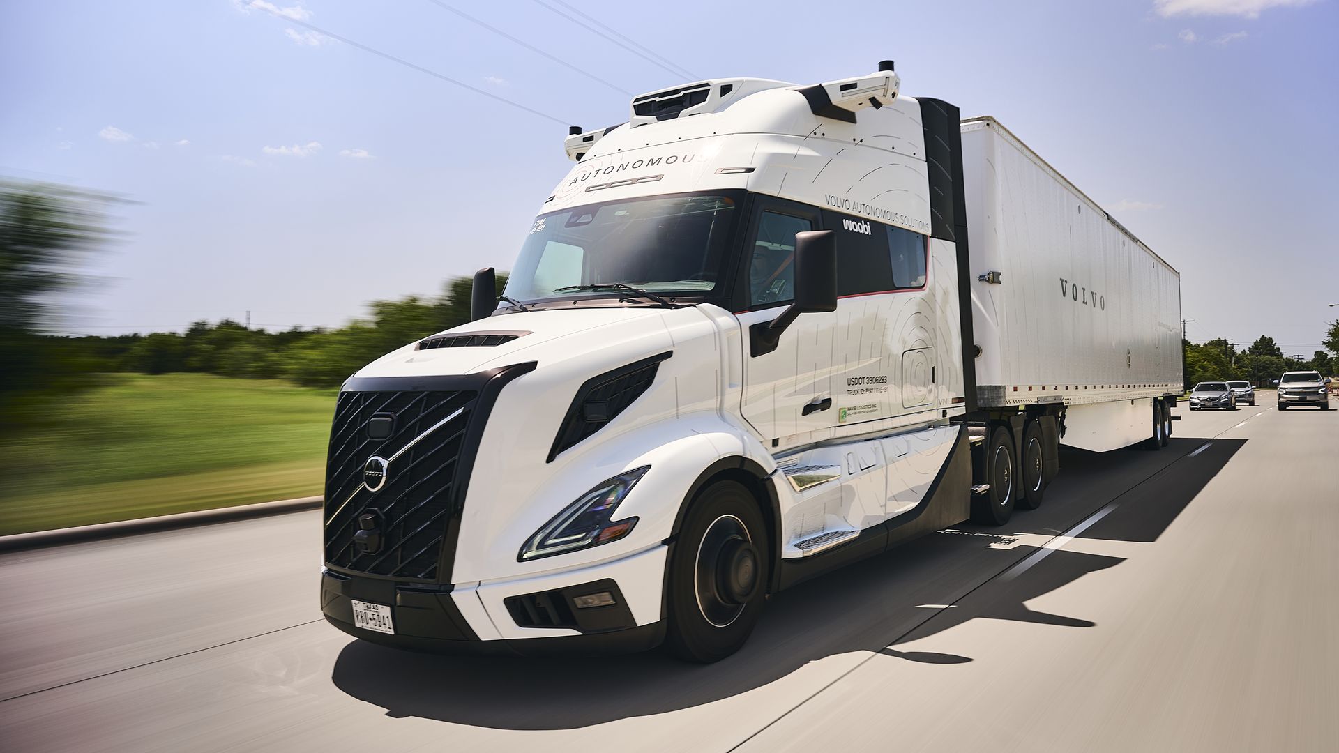 White semi-truck with a long trailer speeds along a highway under a clear blue sky. Volvo branding on the cab and trailer, with the "Autonomous" hinting at self-driving tech; motion blur conveys speed.