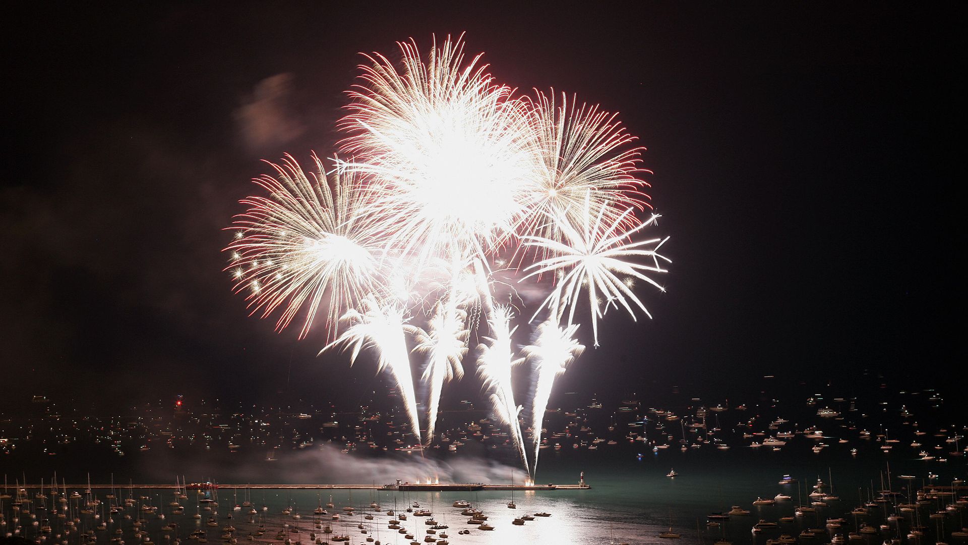 Night scene over a harbor as a large white and red fireworks burst lights the sky. Boats fill the marina, reflections shimmer on the water, and a pier stretches along the foreground.
