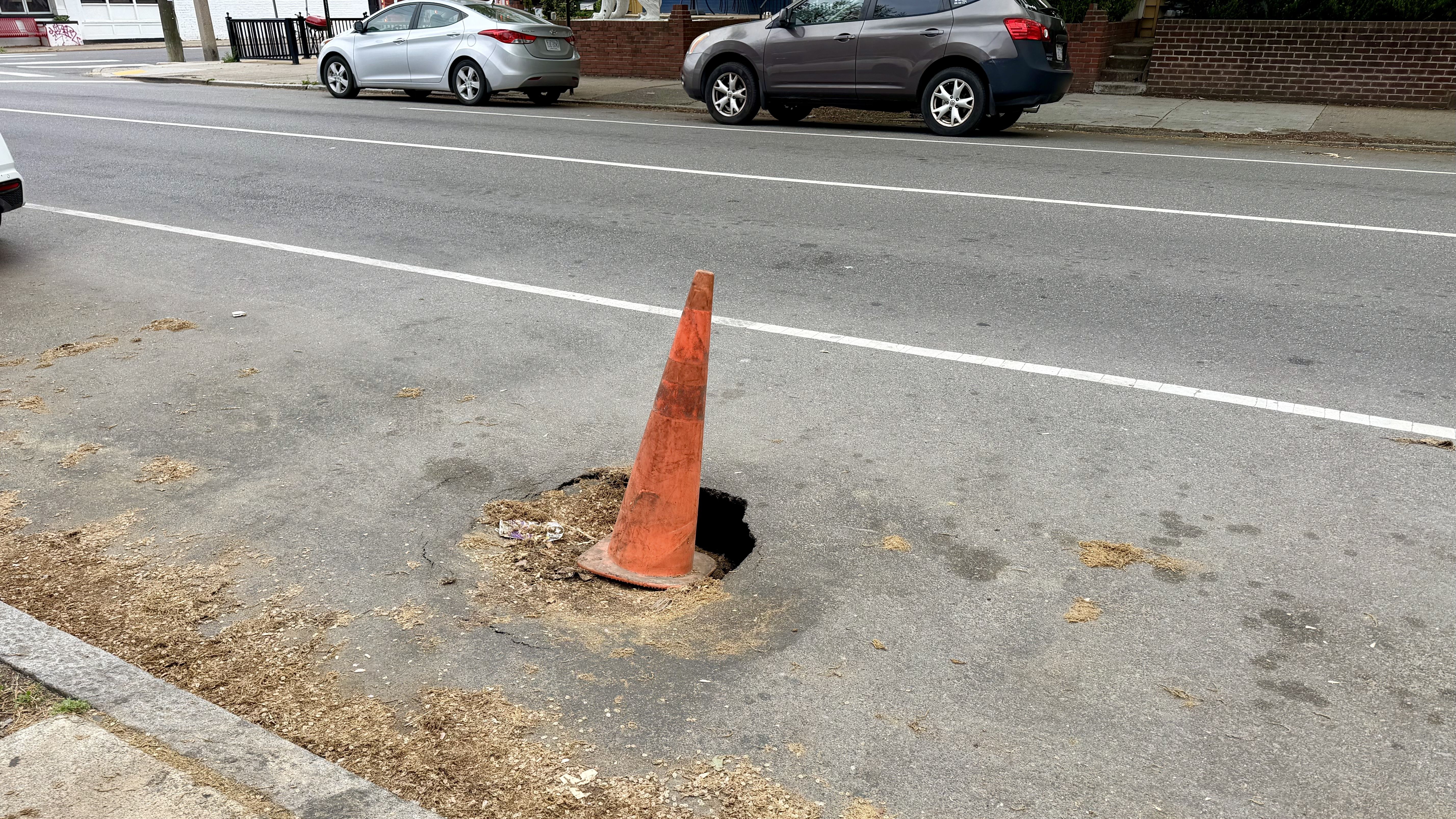 An orange traffic cone sits in a large pothole in a gray street, with white lane lines, parked cars along the curb, a sidewalk, and brick buildings in the background.