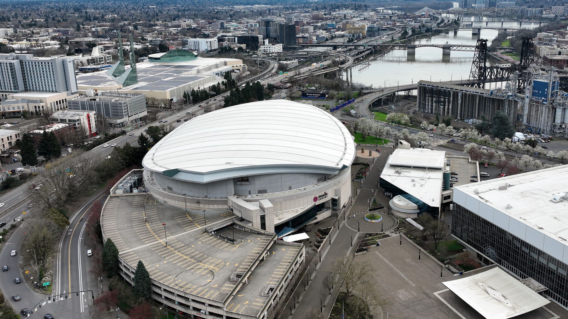 Aerial view of a city along a wide river with several bridges; in the foreground a large white curved-roof building and turquoise glass pyramid-topped structures, plus roads, trees, and mid-rise buildings.