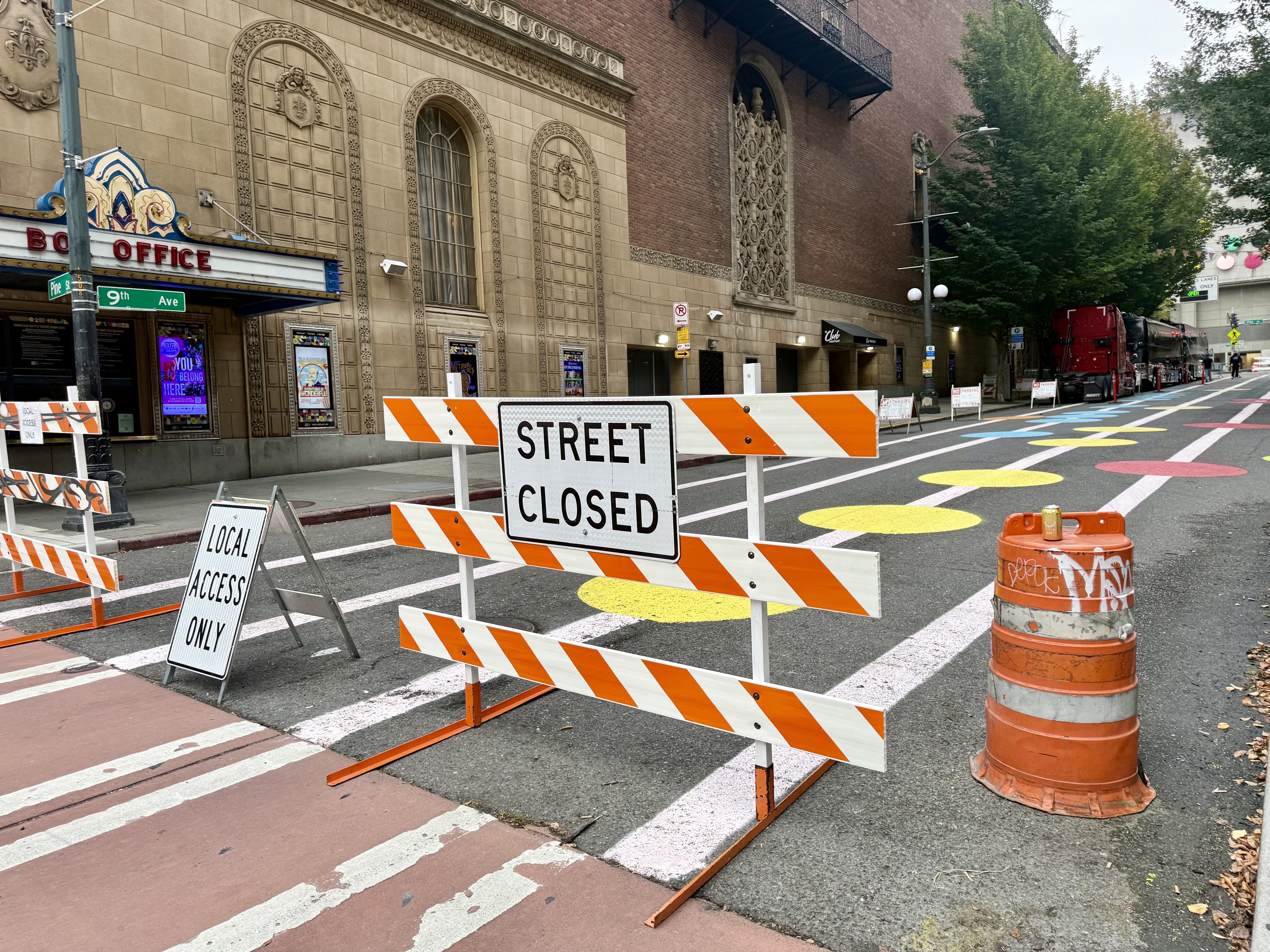 Urban street scene with orange barricades and a Street Closed sign. Ornate beige stone building with tall arched windows, colorful crosswalk circles, and a line of trucks on the right.