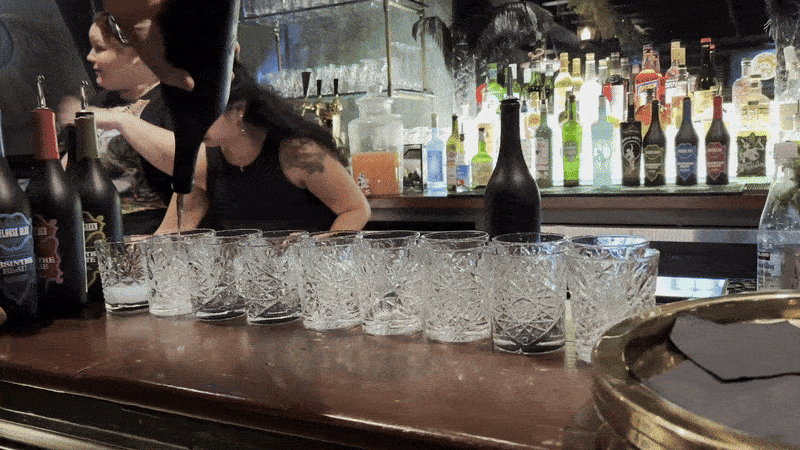 A person turns a bottle of absinthe upside down over glasses lined up on a bar.