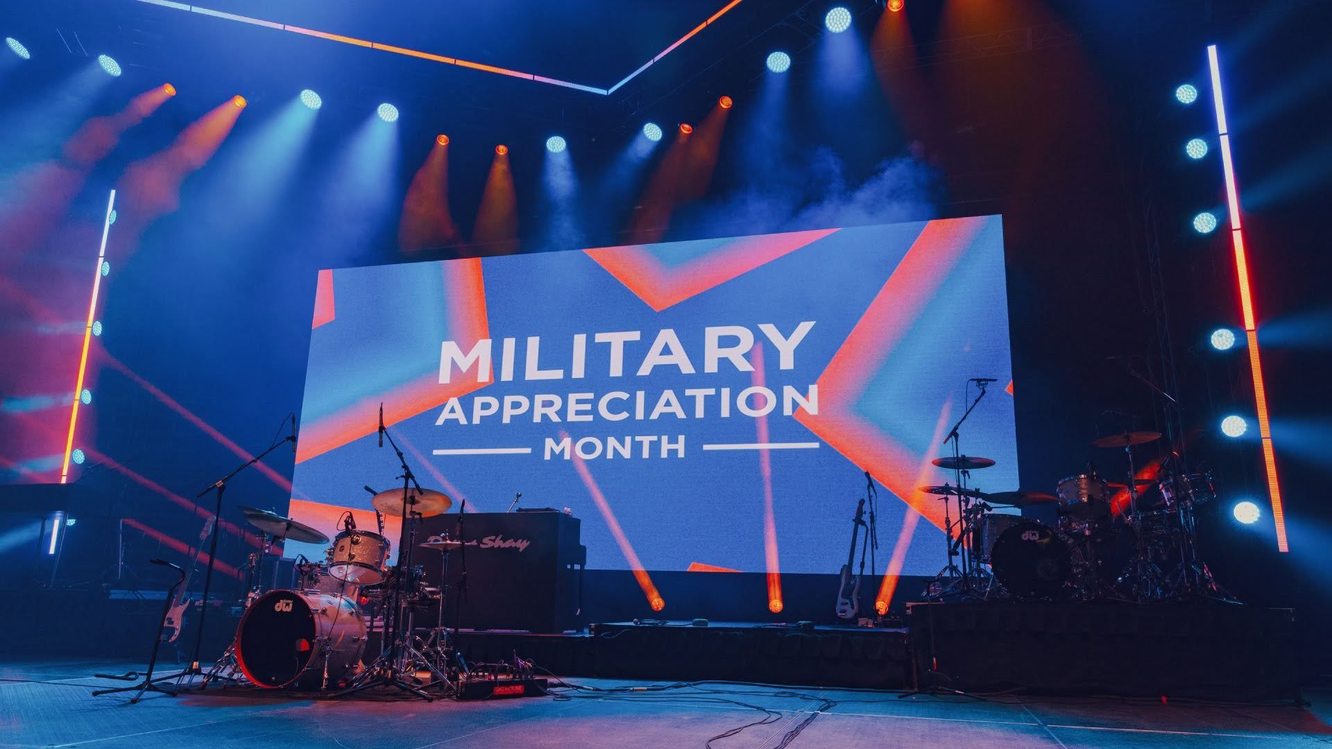 Concert stage with a large LED screen reading "MILITARY APPRECIATION MONTH" in white on a blue and orange geometric backdrop; drums, guitars, and microphones arranged beneath colorful blue and orange lights.