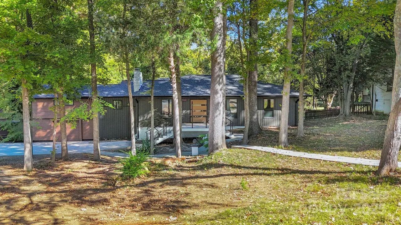 Dark gray single-story house with wood garage doors, surrounded by tall green trees and a lawn with patches of sunlight and shadows.