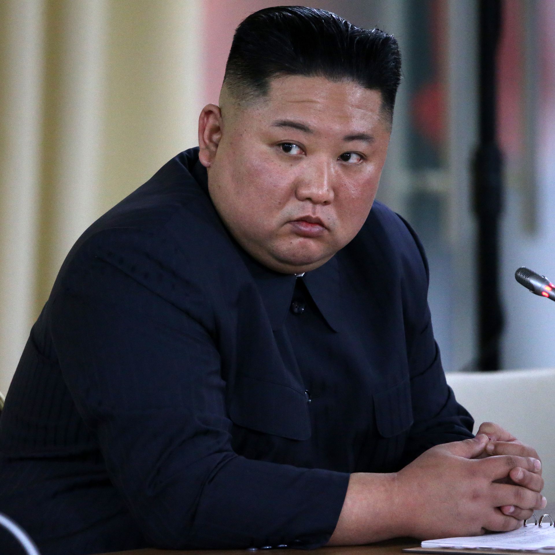 This image shows Kim John Un sitting in front of a microphone and looking to the left.