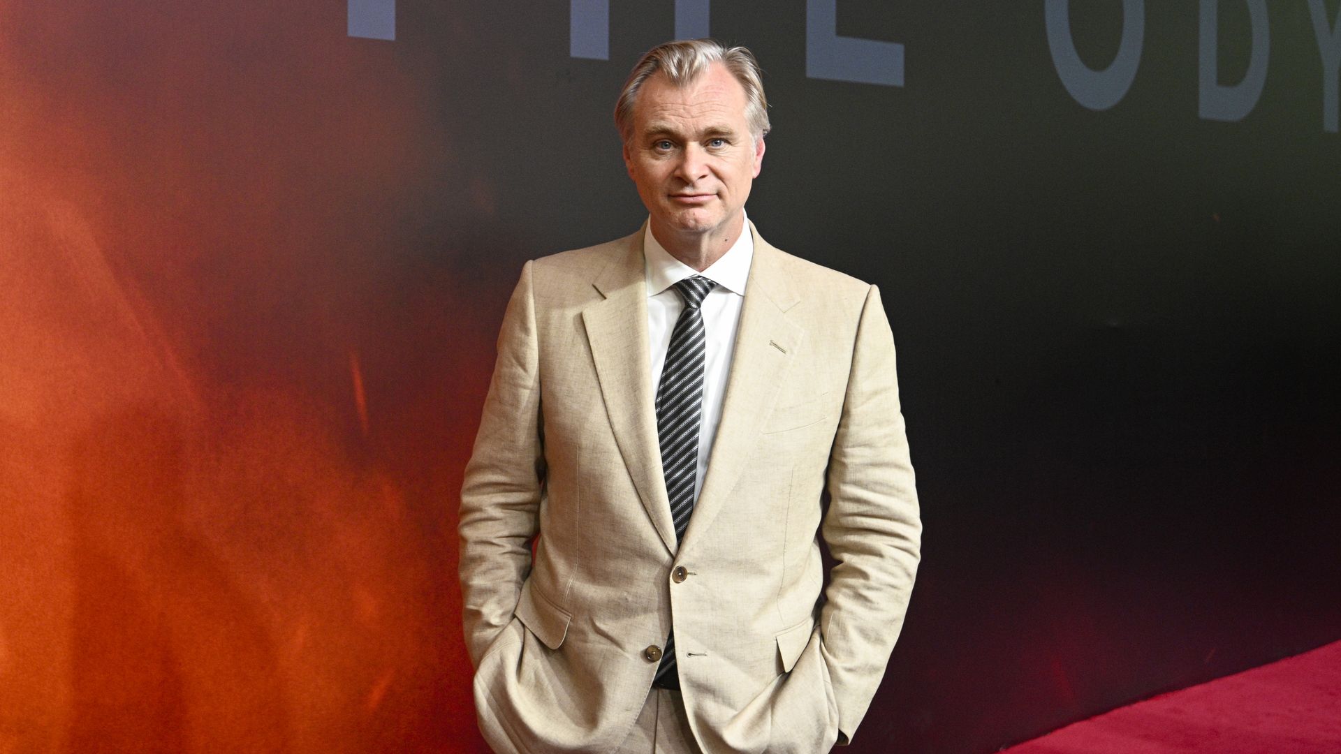 A man in a light beige suit, white shirt, and striped tie stands with hands in pockets, facing the camera with a slight smile, against a red-to-dark backdrop.