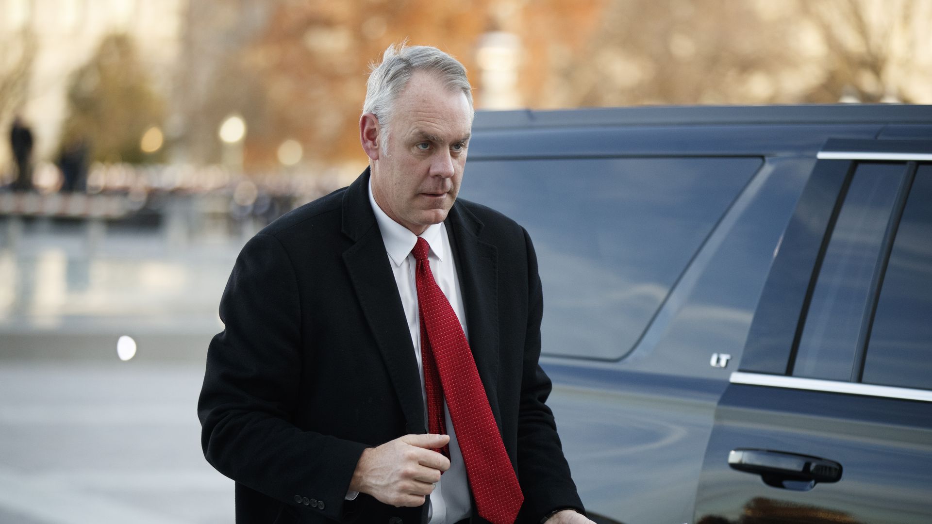 In this image, Ryan Zinke walks out of a black SUV with a red tie.