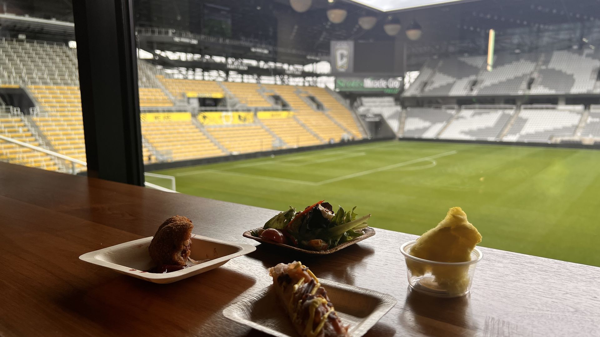 Partial view of stadium seating with yellow and white seats and a soccer field, seen through window behind wooden table holding small plates of food including salad, hot dog, and fried item.