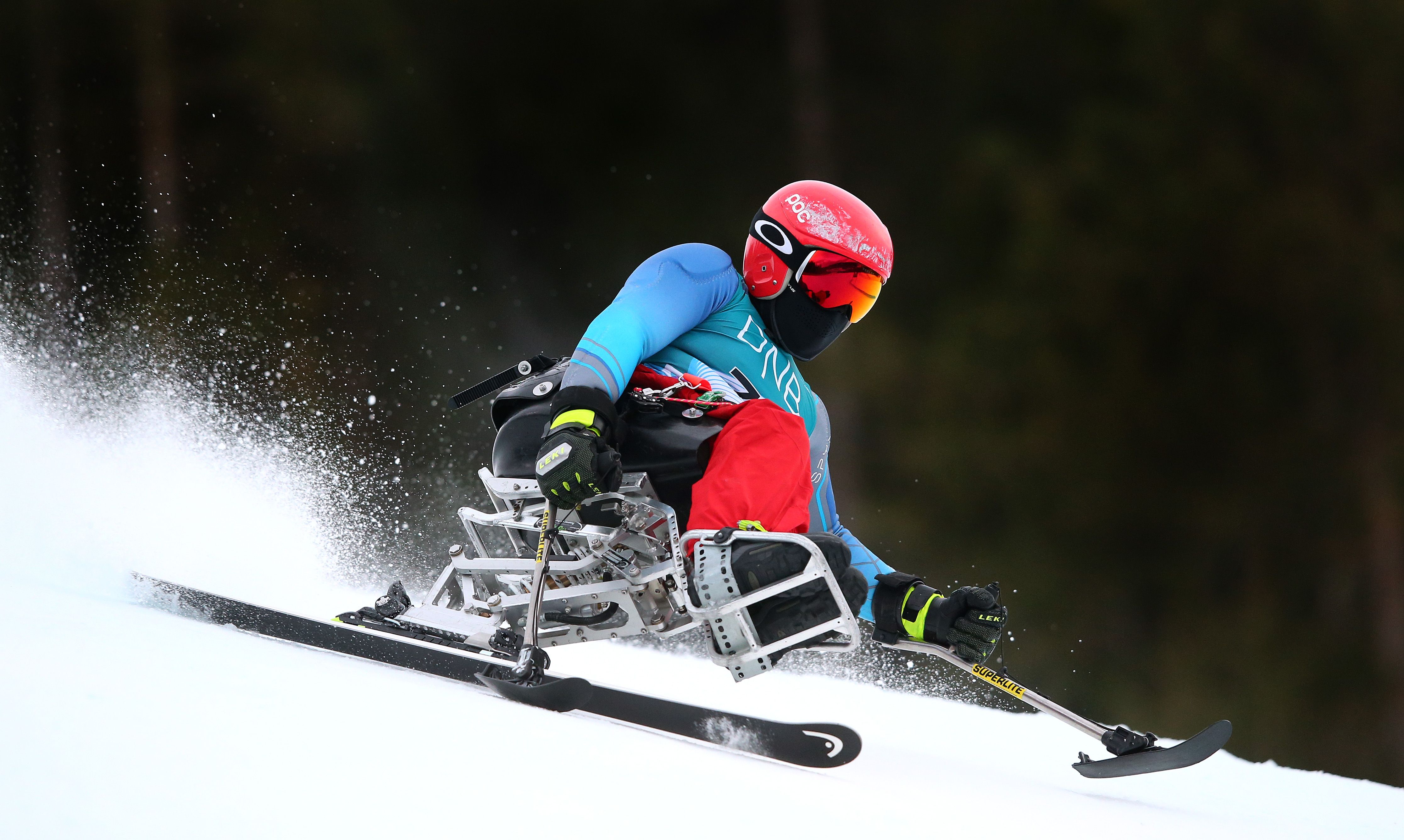 Para alpine skier in red helmet, blue jacket, and red pants racing downhill on snow using a sit-ski with poles, shooting snow spray against a dark forest background.