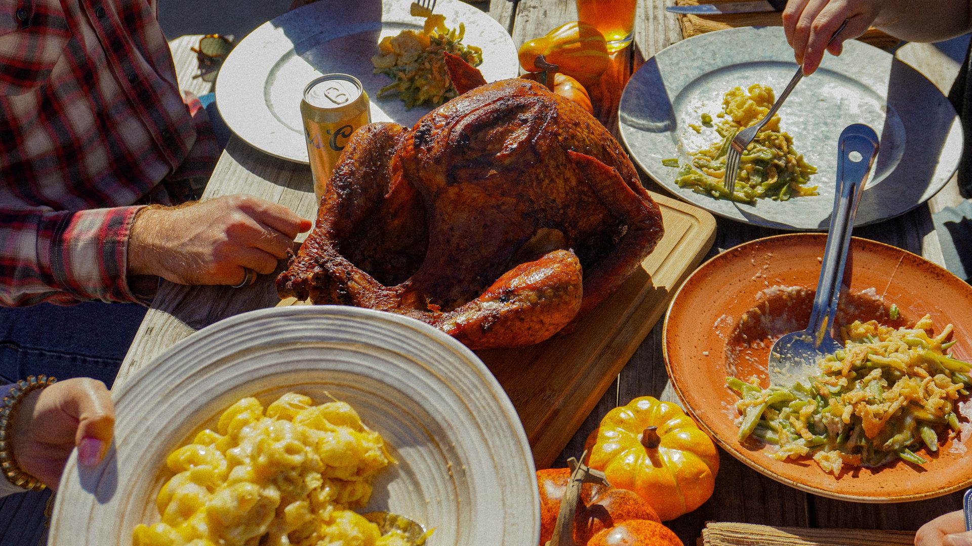 Outdoor Thanksgiving meal with roasted turkey on wooden board, macaroni and cheese, green bean casserole, drinks, and small pumpkins on a rustic wooden table with people eating.