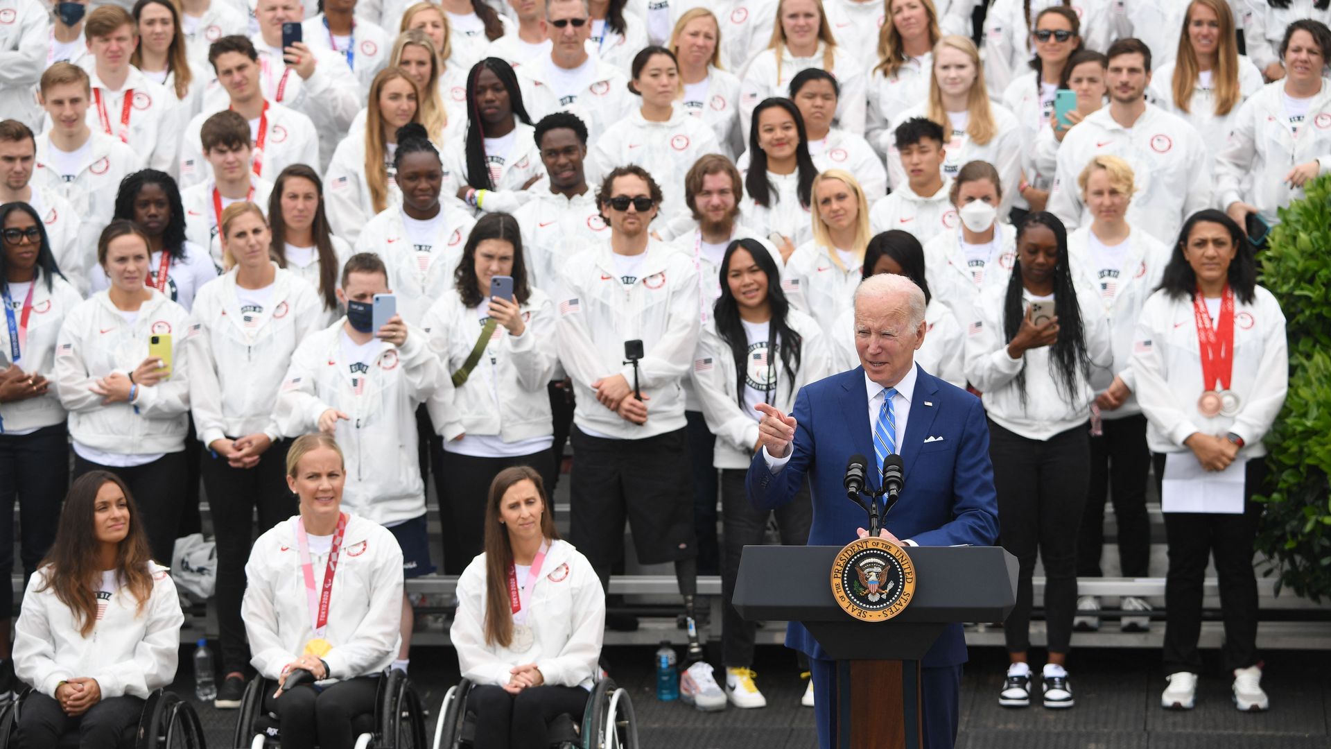 Photo of Joe Biden speaking from a podium with rows of people wearing white standing behind him and seated at wheelchairs beside him