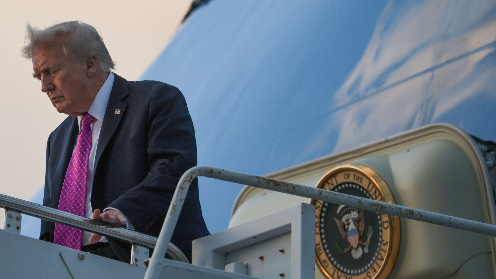 Trump — wearing a dark suit, a pink tie and a white collared shirt — touches his hand to a railing as he exits Air Force One.