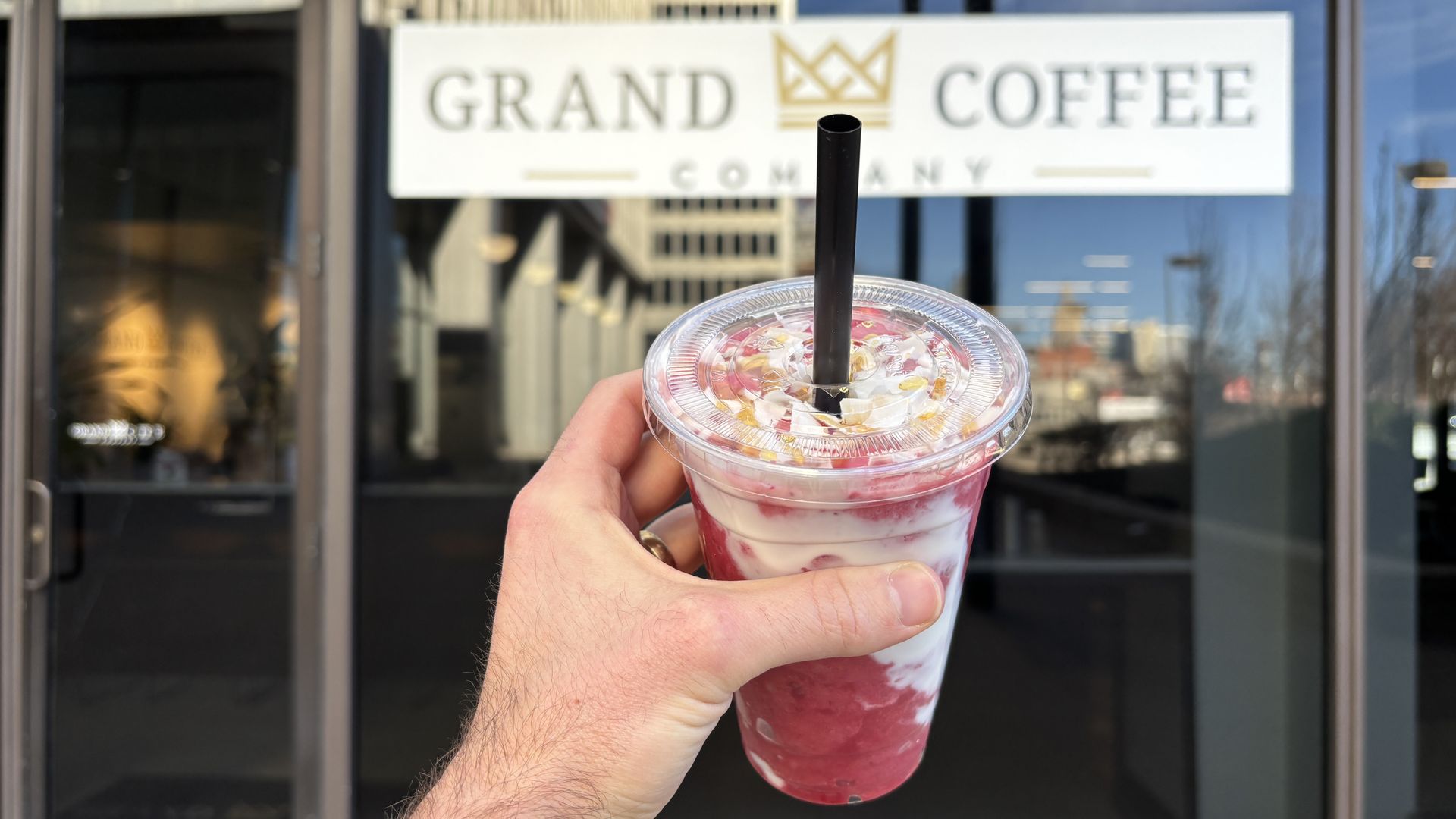 Hand holding a clear cup with a black straw containing a white and red layered drink topped with whipped cream and crushed nuts, in front of a glass door with a "GRAND COFFEE COMPANY" sign.