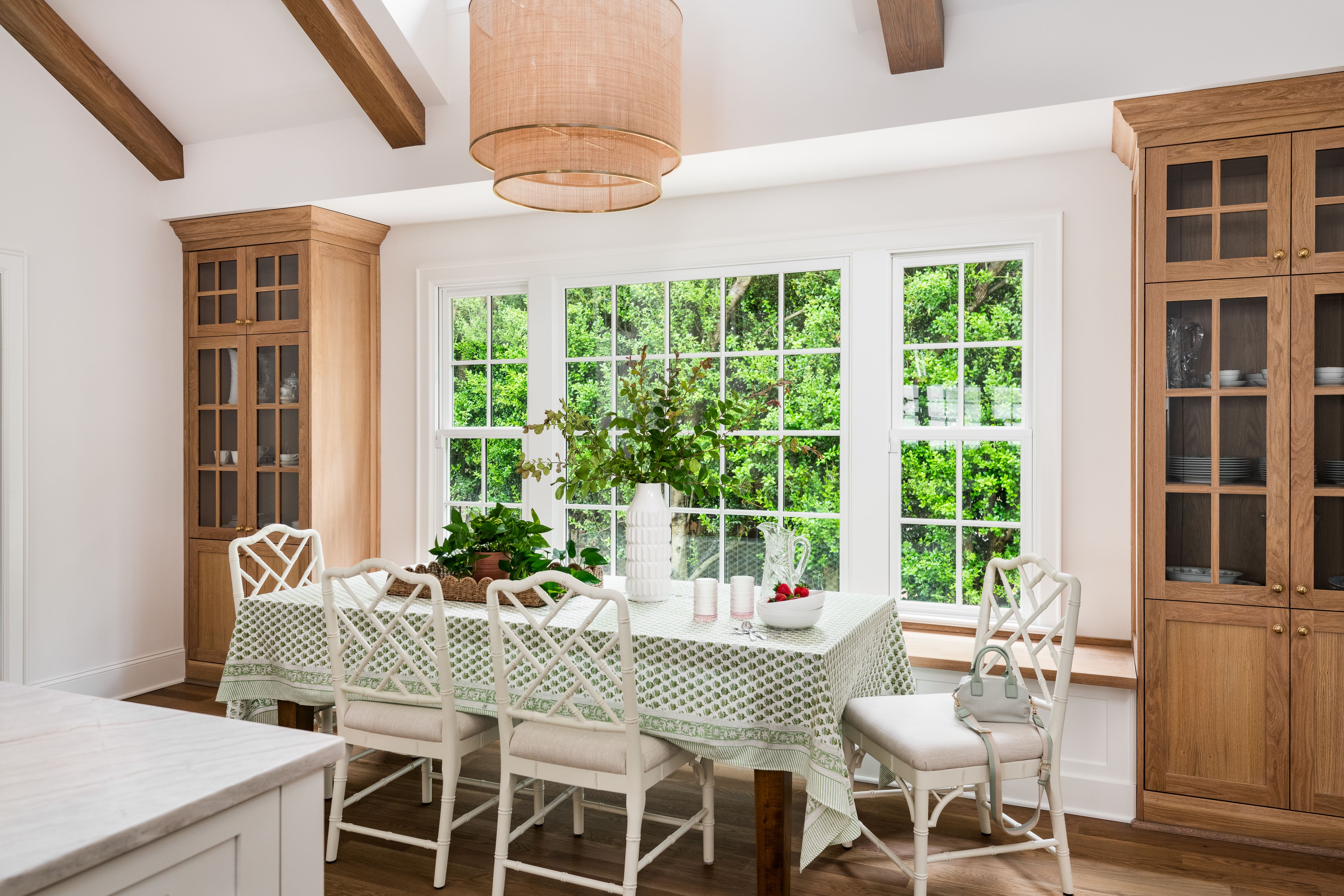 Bright dining room with white-framed windows showing green trees outside, white chairs with cushions, wooden cabinets, green patterned tablecloth, and a large beige pendant light.