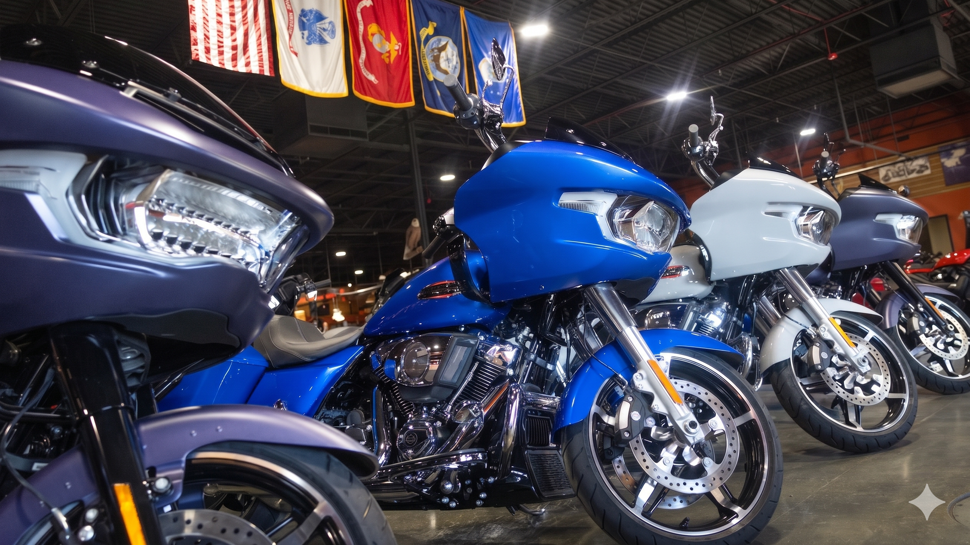 Inside a motorcycle showroom with a row of blue and gray bikes, chrome engines and bright headlights; hanging flags including the U.S. flag sway under industrial lighting.