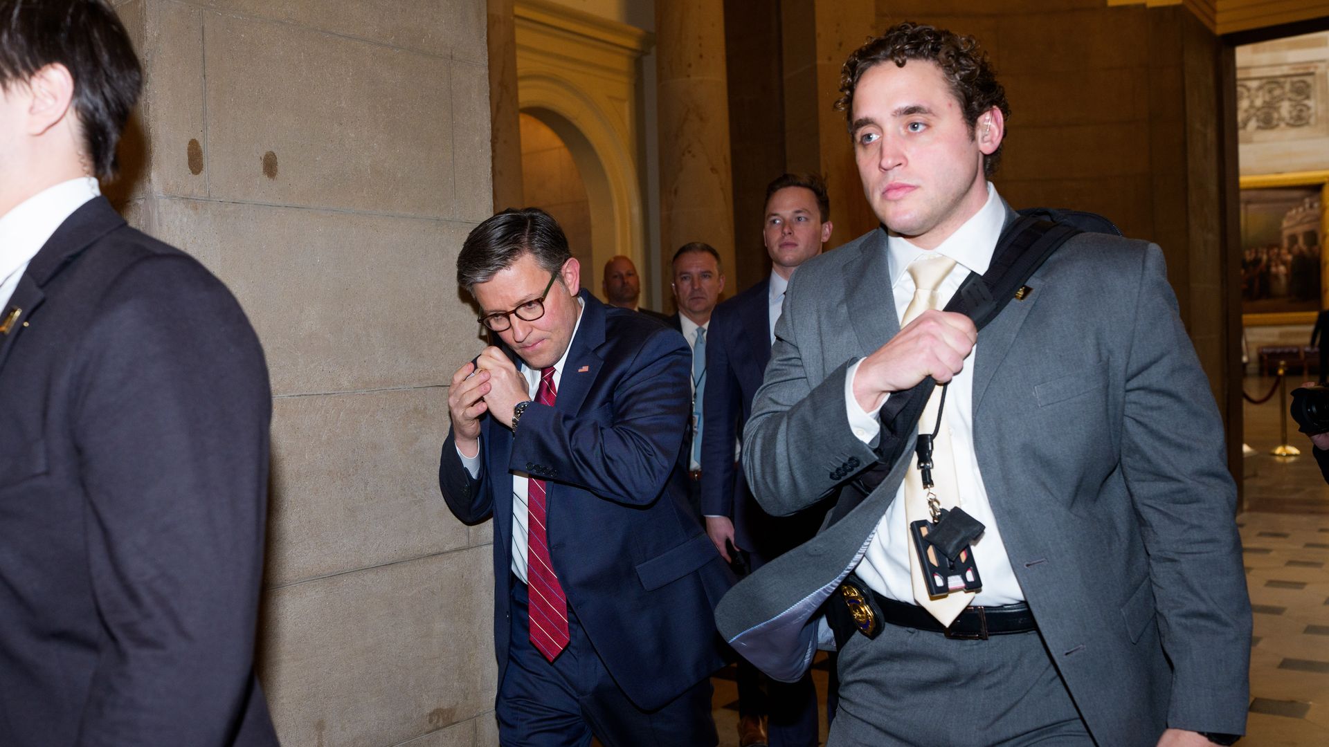 Men in suits walking through a hallway with stone walls and archways; one man in a gray suit carries a bag and keys, another in a navy suit adjusts his earpiece.