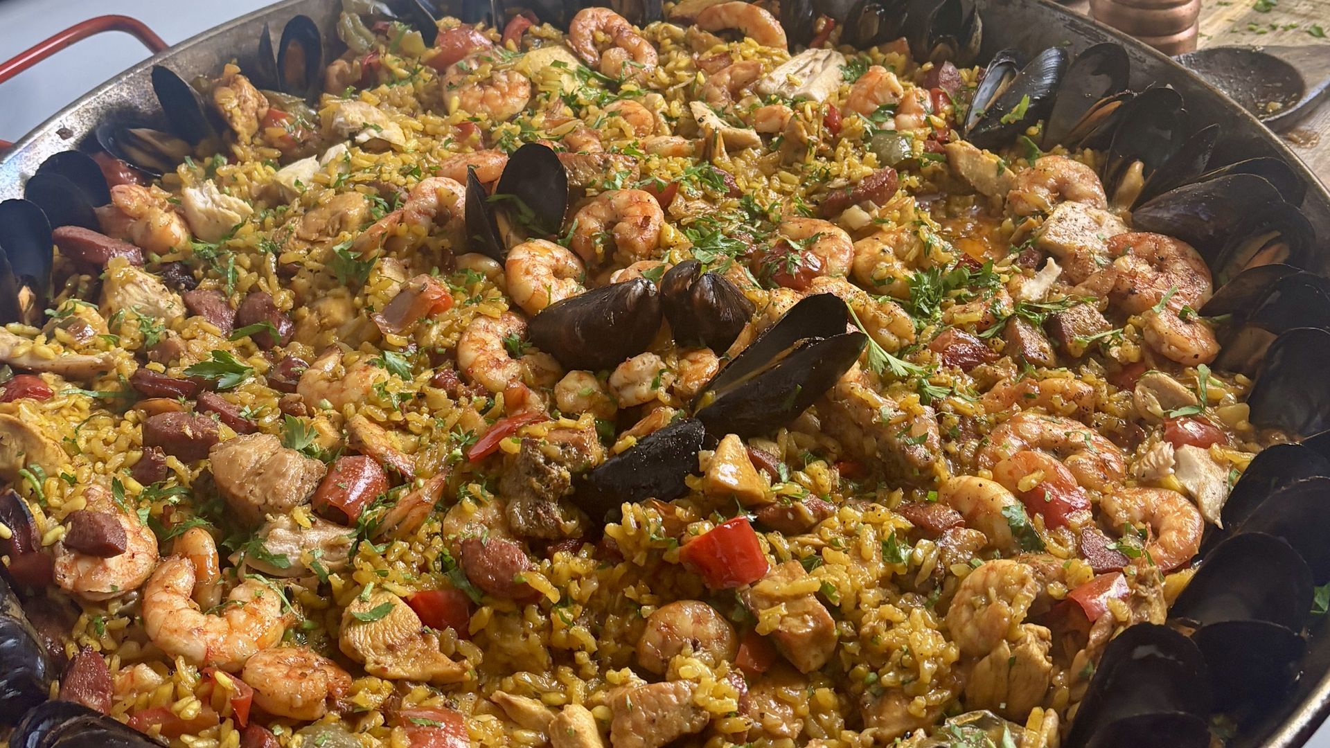 Large skillet of paella with shrimp, mussels, peppers, and peas on a stove. Background shows olive oil bottle, parsley, a can, and assorted kitchen items on the counter.