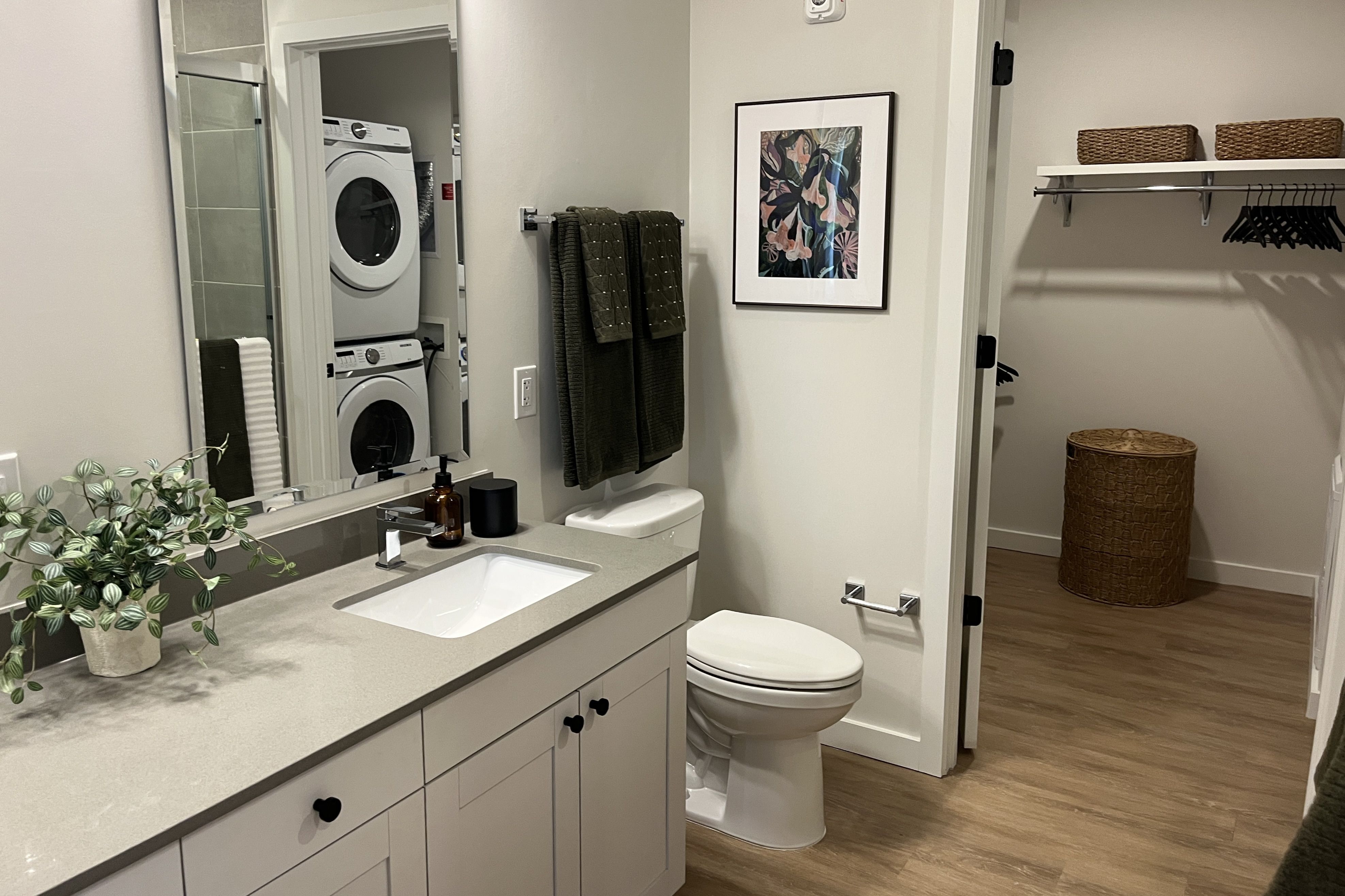Modern bathroom with light gray vanity, white sink and chrome faucet, potted plant, dark towels, framed floral art, open closet with wicker baskets, and a stacked washer/dryer seen in the mirror.