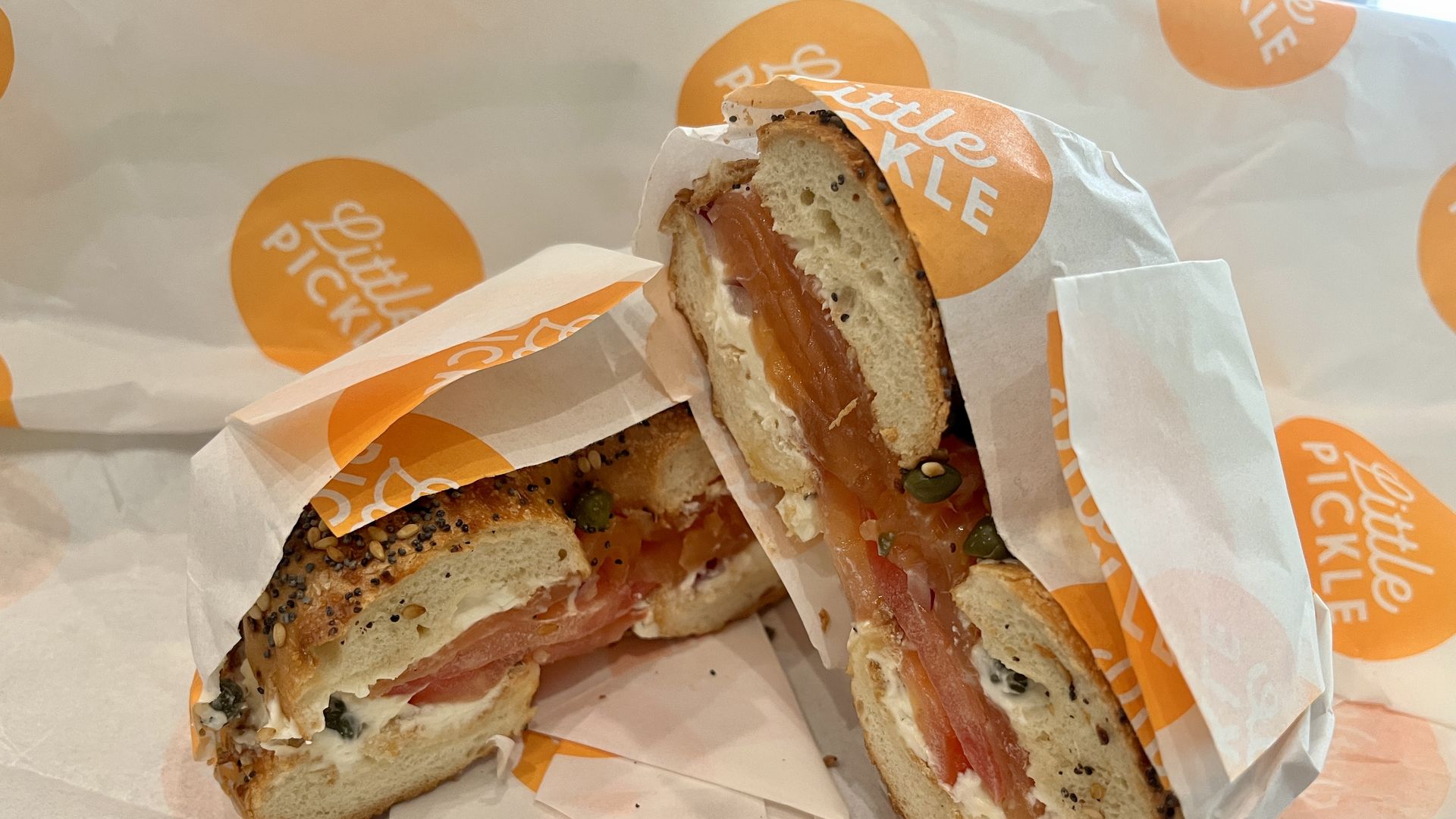 A sliced everything bagel sandwich wrapped in white and orange "Little Pickle" paper, filled with cream cheese, smoked salmon, tomato, and capers.