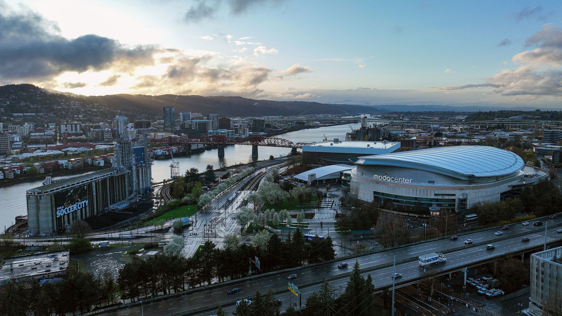 Aerial view of a city at sunset: the Moda Center with a curved roof on the riverfront, a bridge crossing the water, a large "SoccerCity" structure, highways, trees, and distant hills.