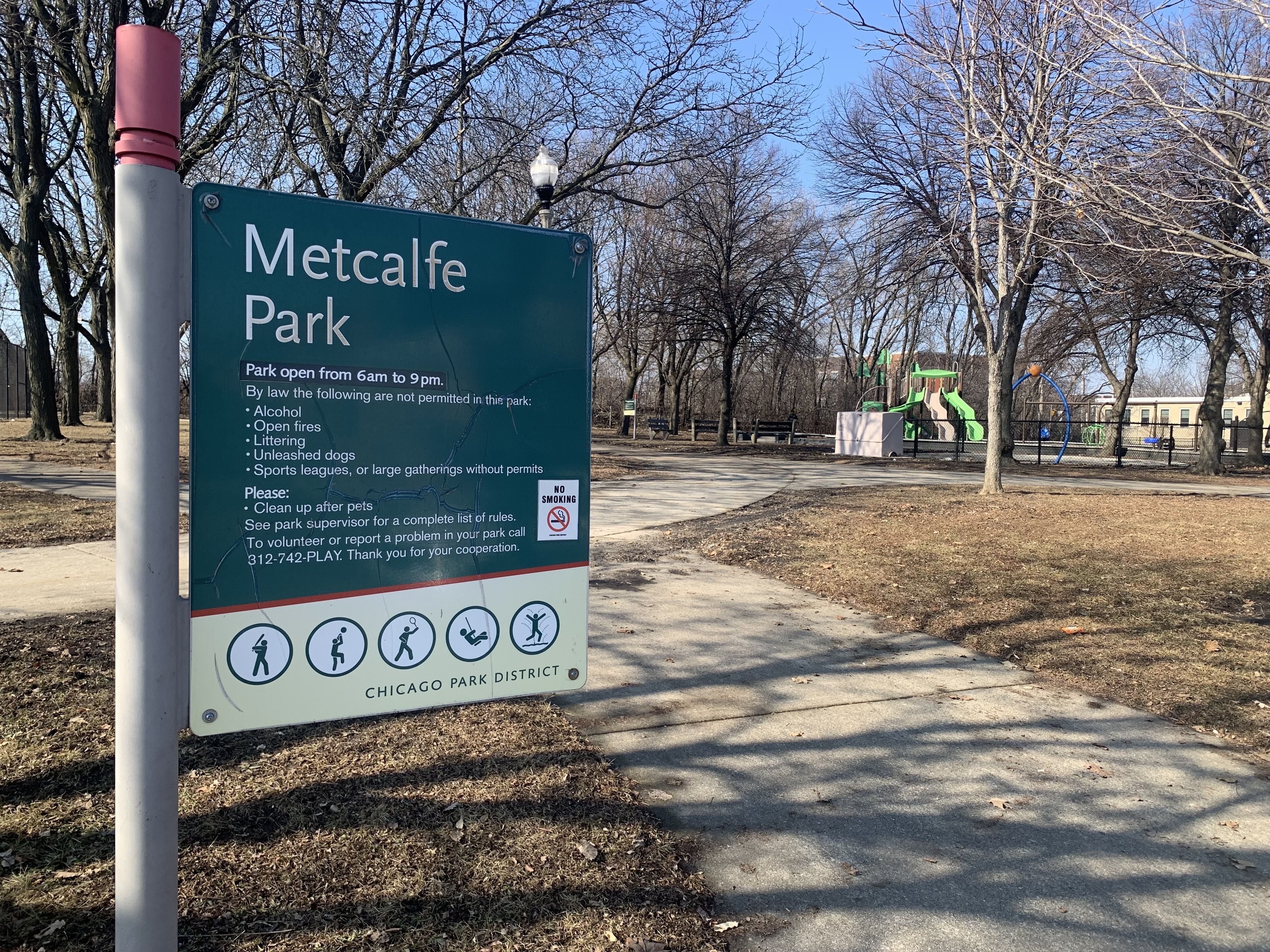 Sign for Metcalfe Park in Chicago Park District with rules including no alcohol, open fires, littering, unleashed dogs, and large gatherings. Playground with green slides in background.