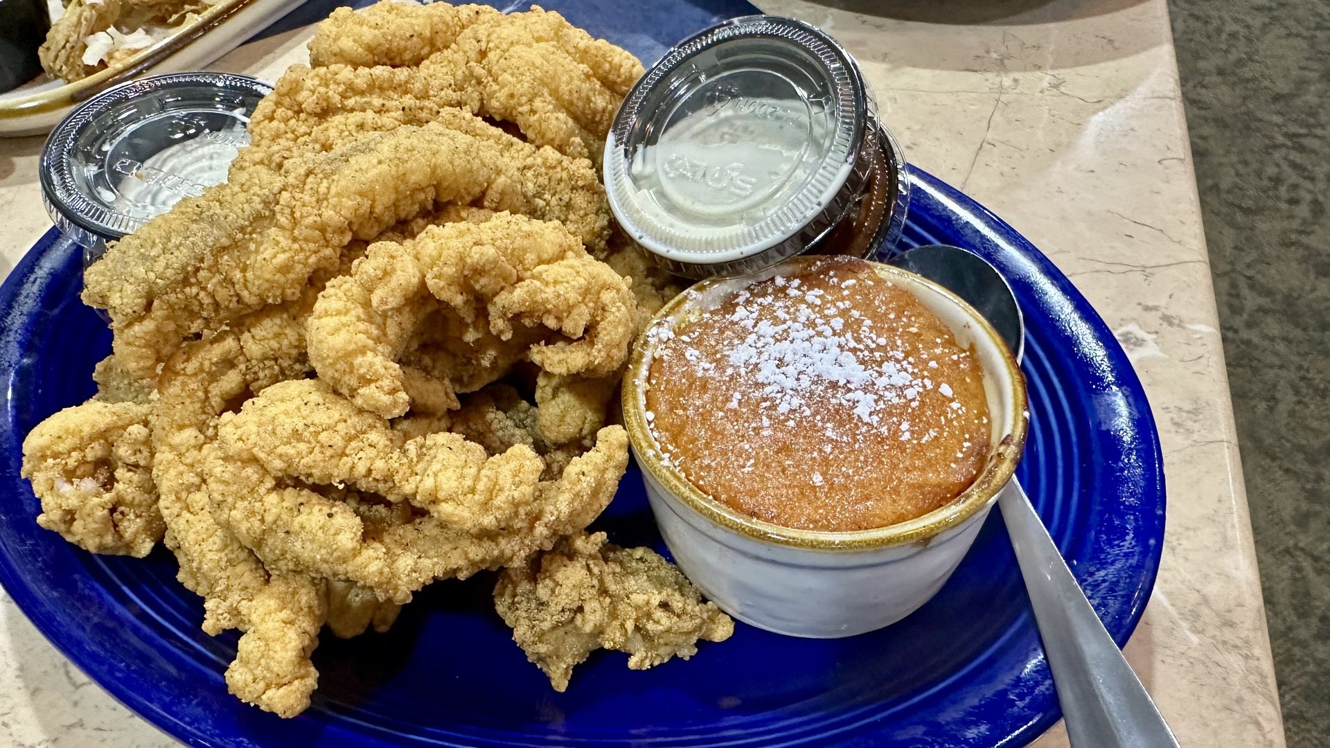 Blue plate with crispy fried catfish strips, small bowl of carrot soufflé topped with powdered sugar, two plastic containers of white dipping sauce, and a silver spoon on a light-colored table.