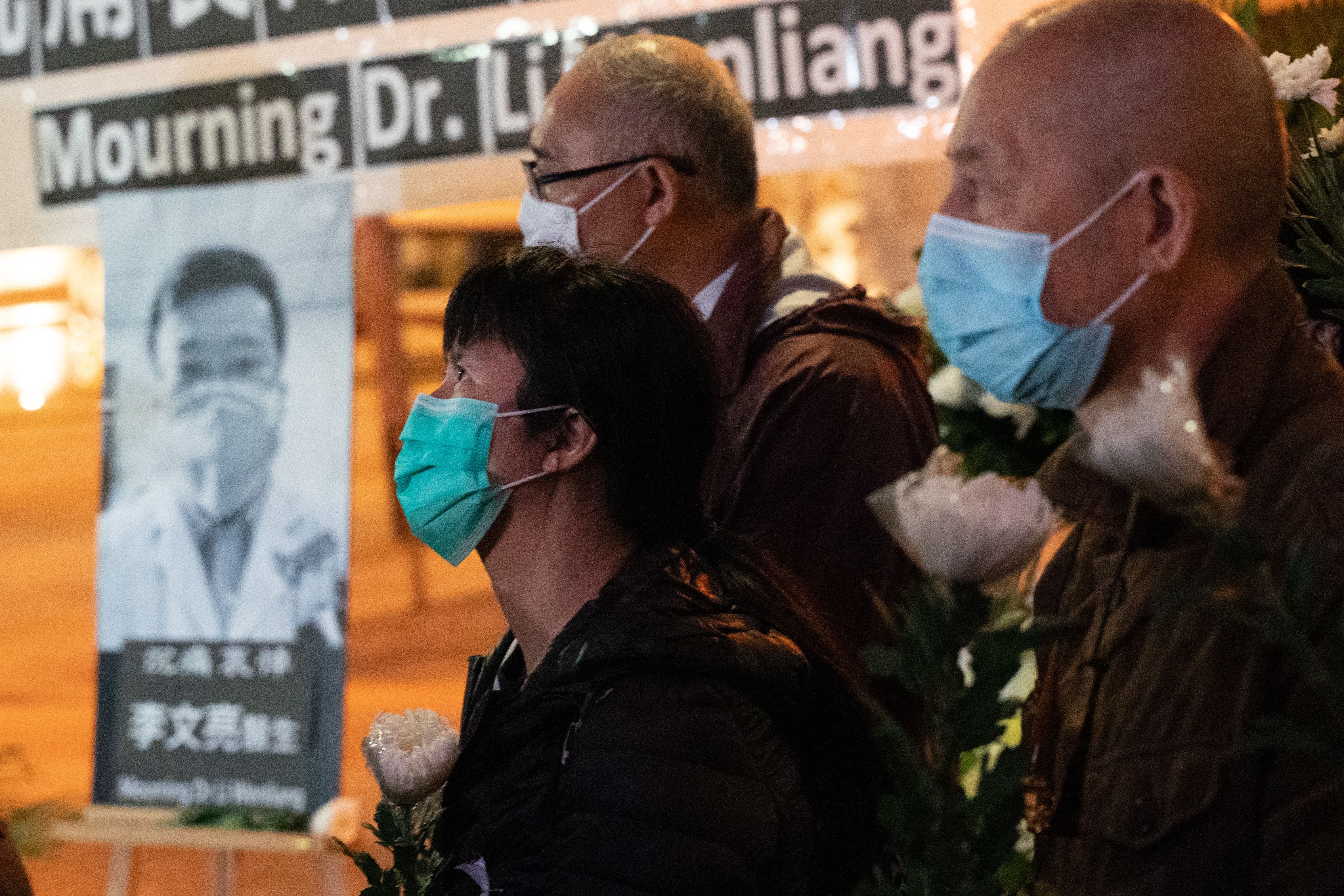 In this image, people with protective face masks stand next to a photo of Dr. Li and a sign that says "Mourning Dr. Li" 
