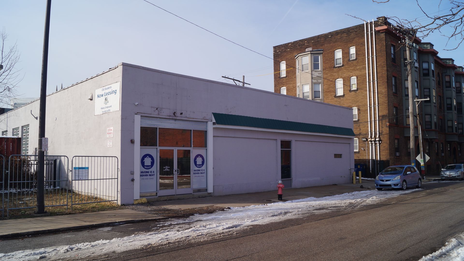 White, single-story building with signs reading "Housing is a Human Right" and a "Now Leasing" banner, a purple car parked on the street, some snow on the ground, and taller brick buildings in the background.