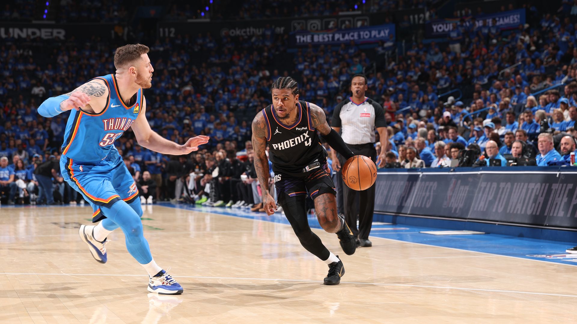 A basketball player in a black uniform with "Phoenix" and the number 4 on the front of his jersey drives with the ball against a defender in a light blue uniform with "Thunder" on the front of the jersey, in a packed arena. 