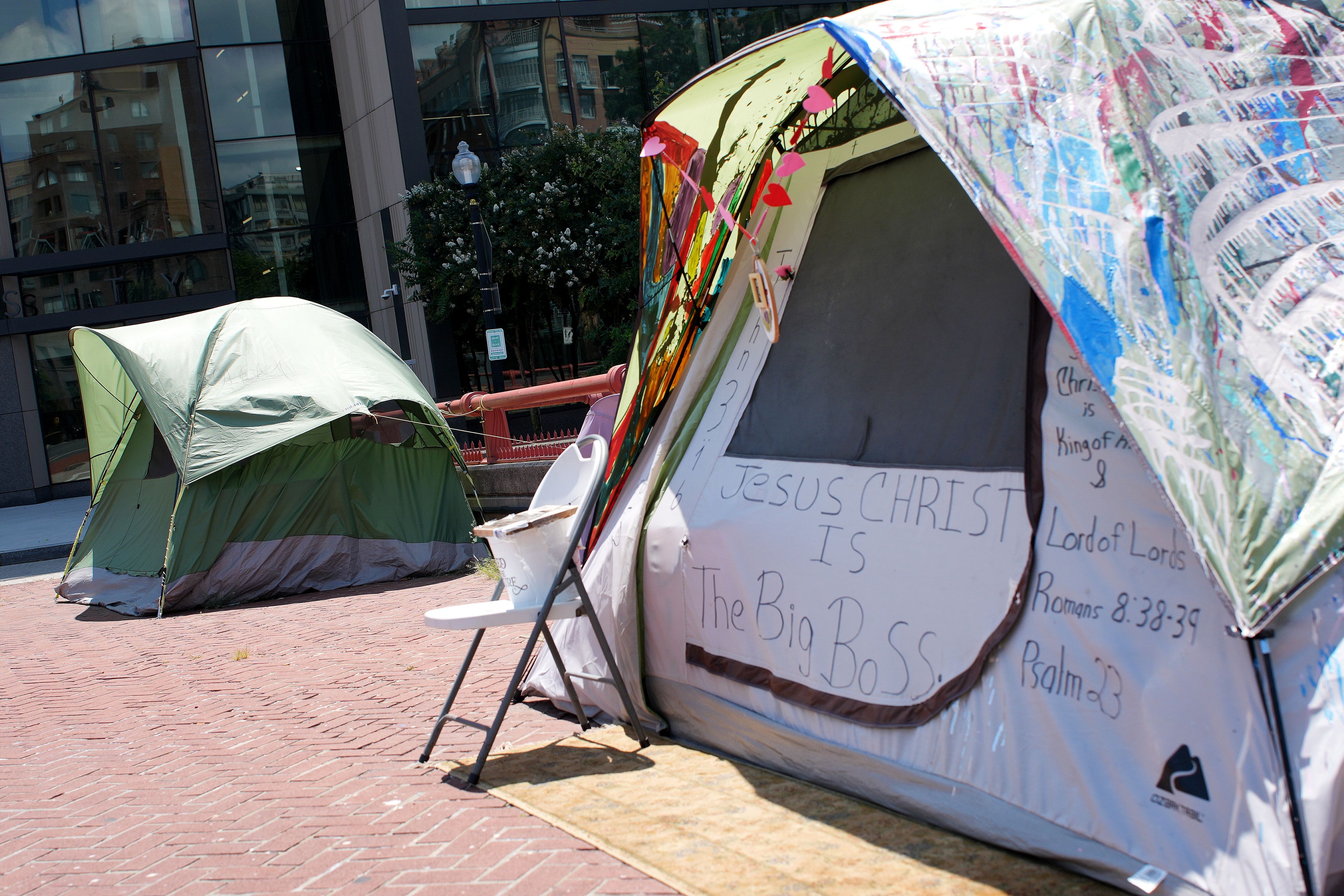 Two tents set on a brick pavement in an urban area; one tent is green, the other is decorated with colorful paint and features signs saying "Jesus CHRIST is The Big Boss" and biblical references.
