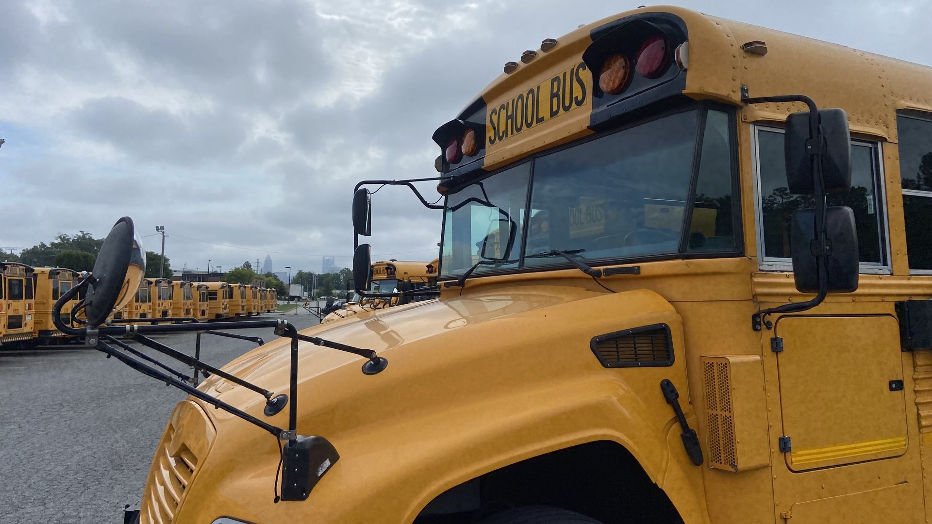 Close-up of a yellow school bus front and side, with several other yellow school buses parked in rows under a cloudy sky.