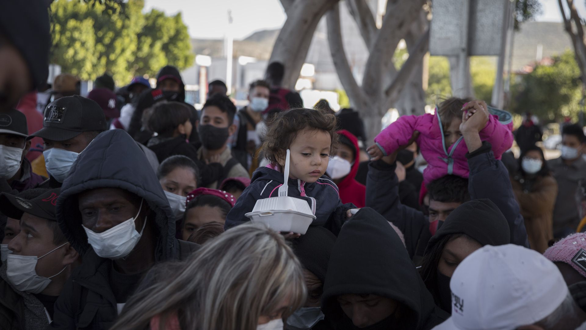 Children are carried on shoulders as scores of asylum seekers wait for a meal and a drink at the "El Chaparral" border crossing