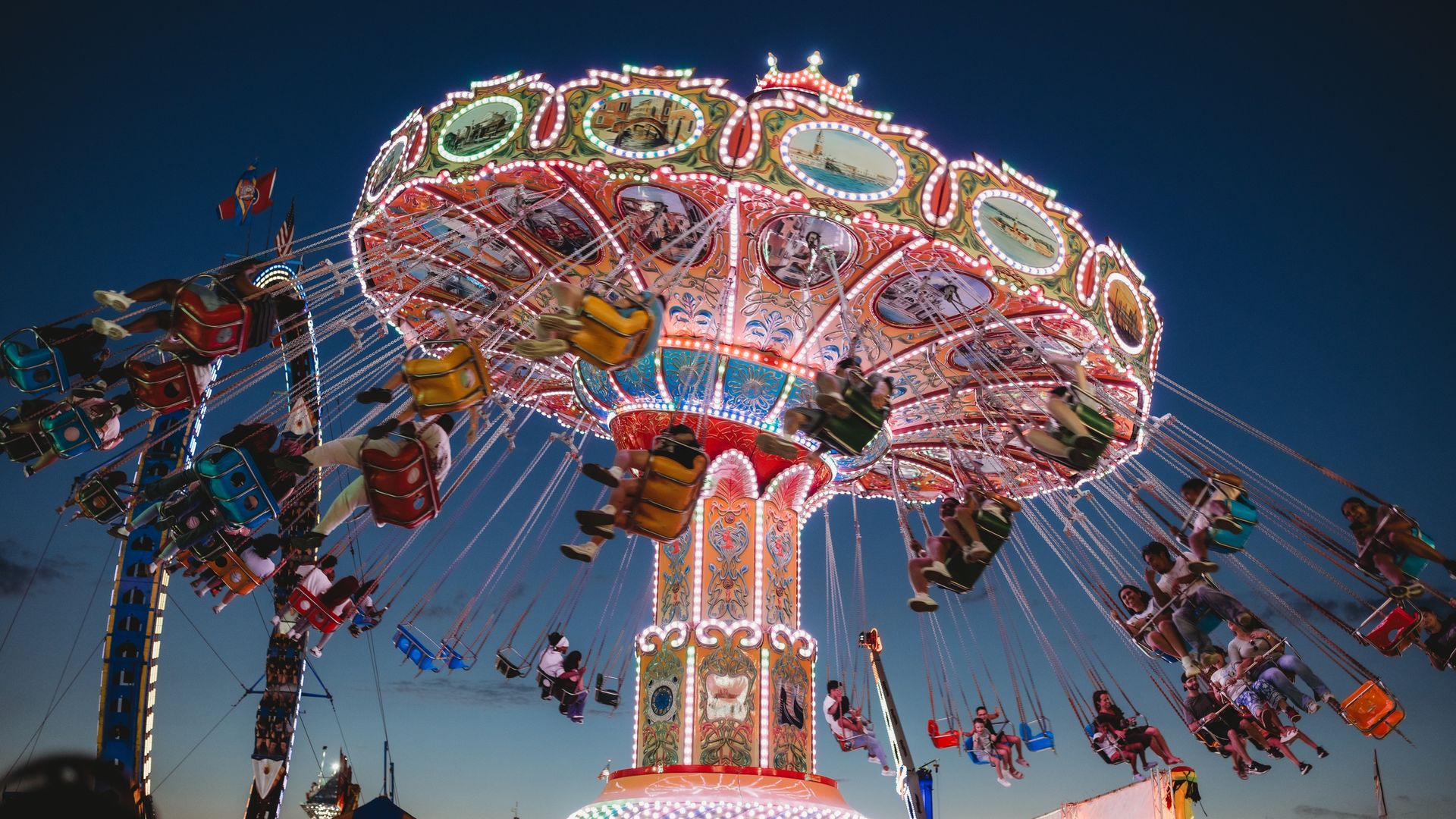 Carnival swing ride at dusk with bright multicolored lights and ornate panels, riders in colorful chairs spinning against a deep blue sky.