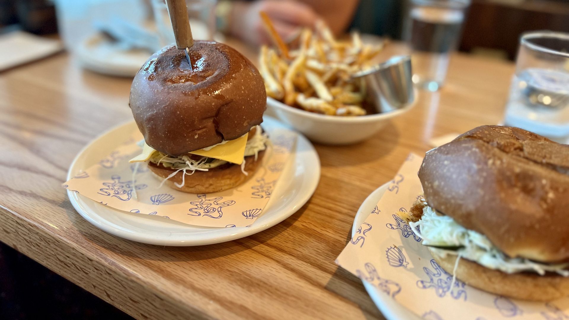 Two sandwiches on white plates with printed paper, one with a wooden stick, and a bowl of fries with sauce on a wooden table in a casual dining setting.