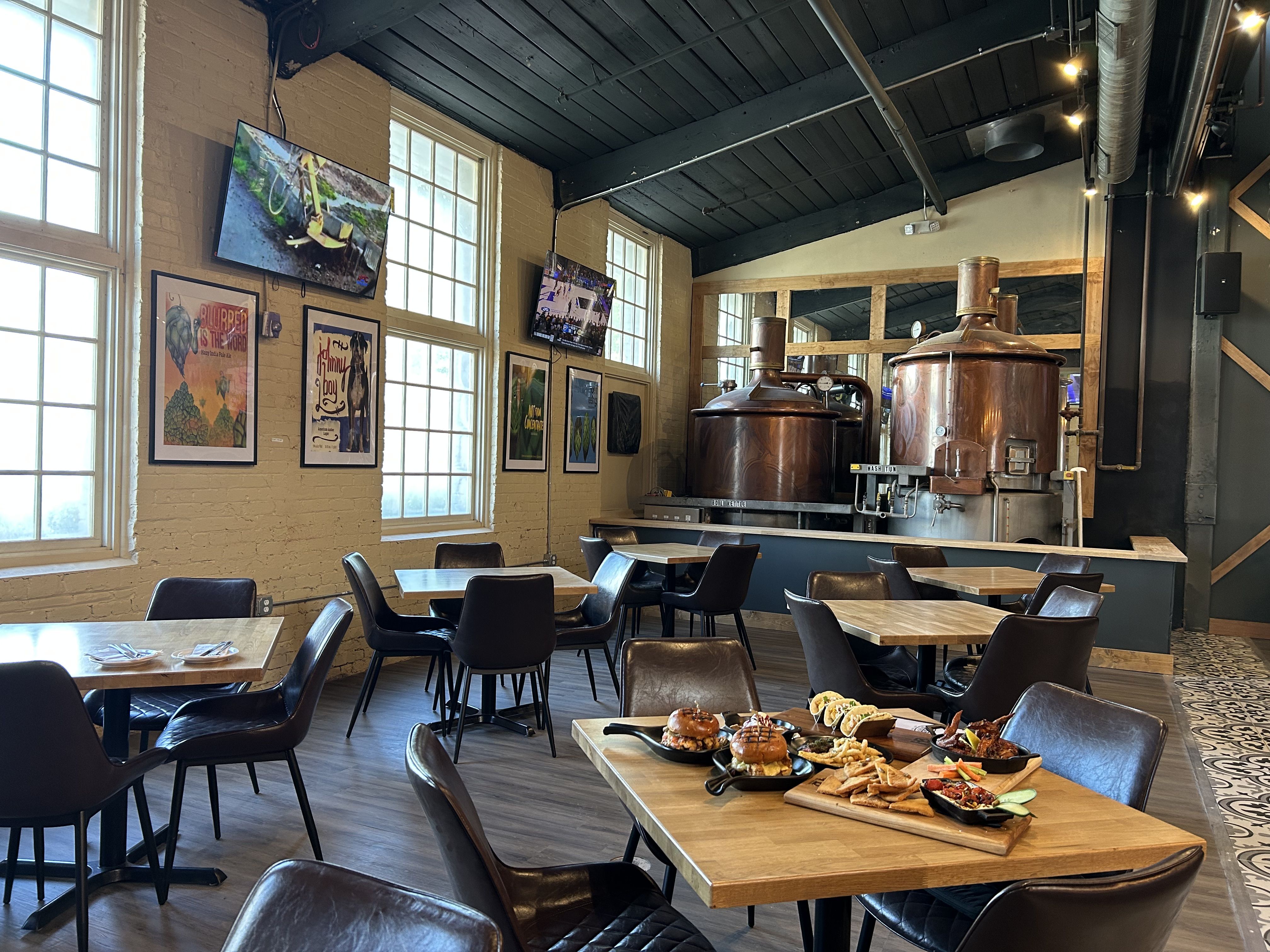 Heist Brewery. Industrial cafe with brick walls, tall windows, and a dark ceiling. Copper brewing kettles line the right, string lights, posters, and TVs. Wooden tables fill the space; a food platter sits in front.