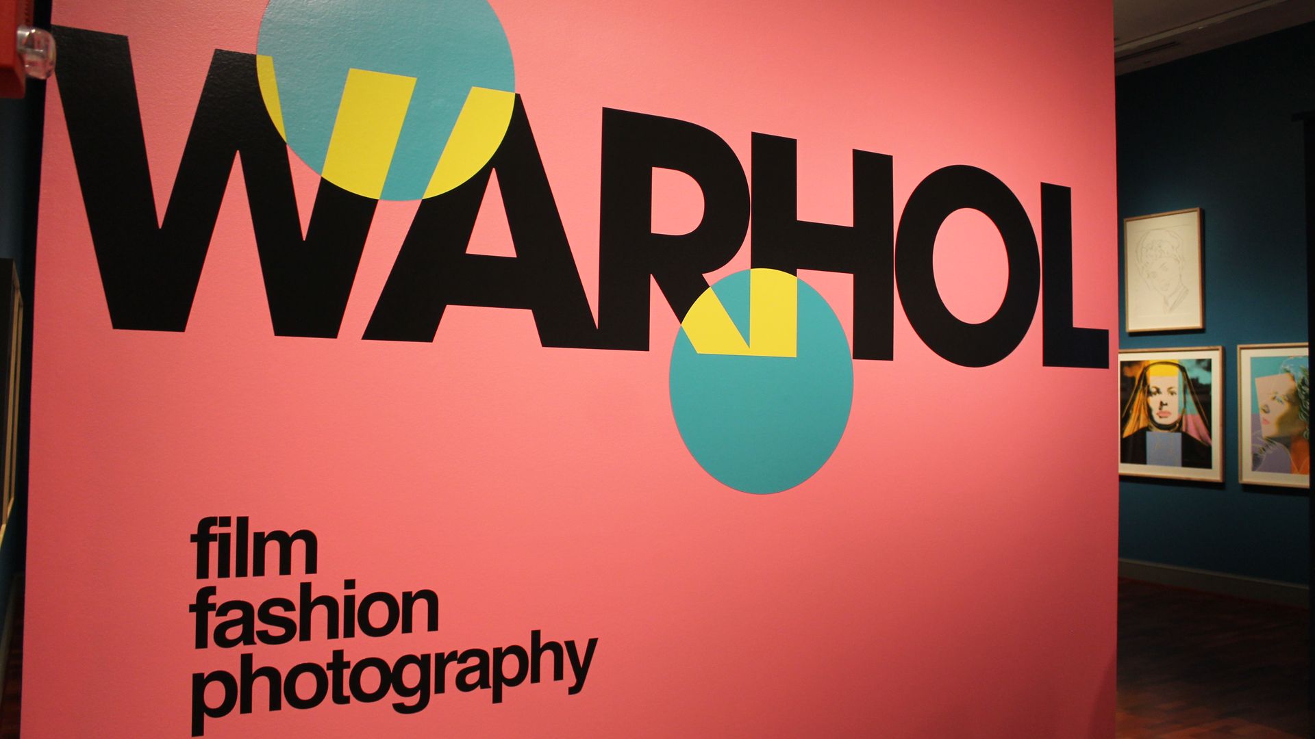 Pink wall with oversized black letters spelling WARHOL, partly obscured by teal and yellow circles; lower left reads film fashion photography; blue gallery wall on the right with framed portraits.