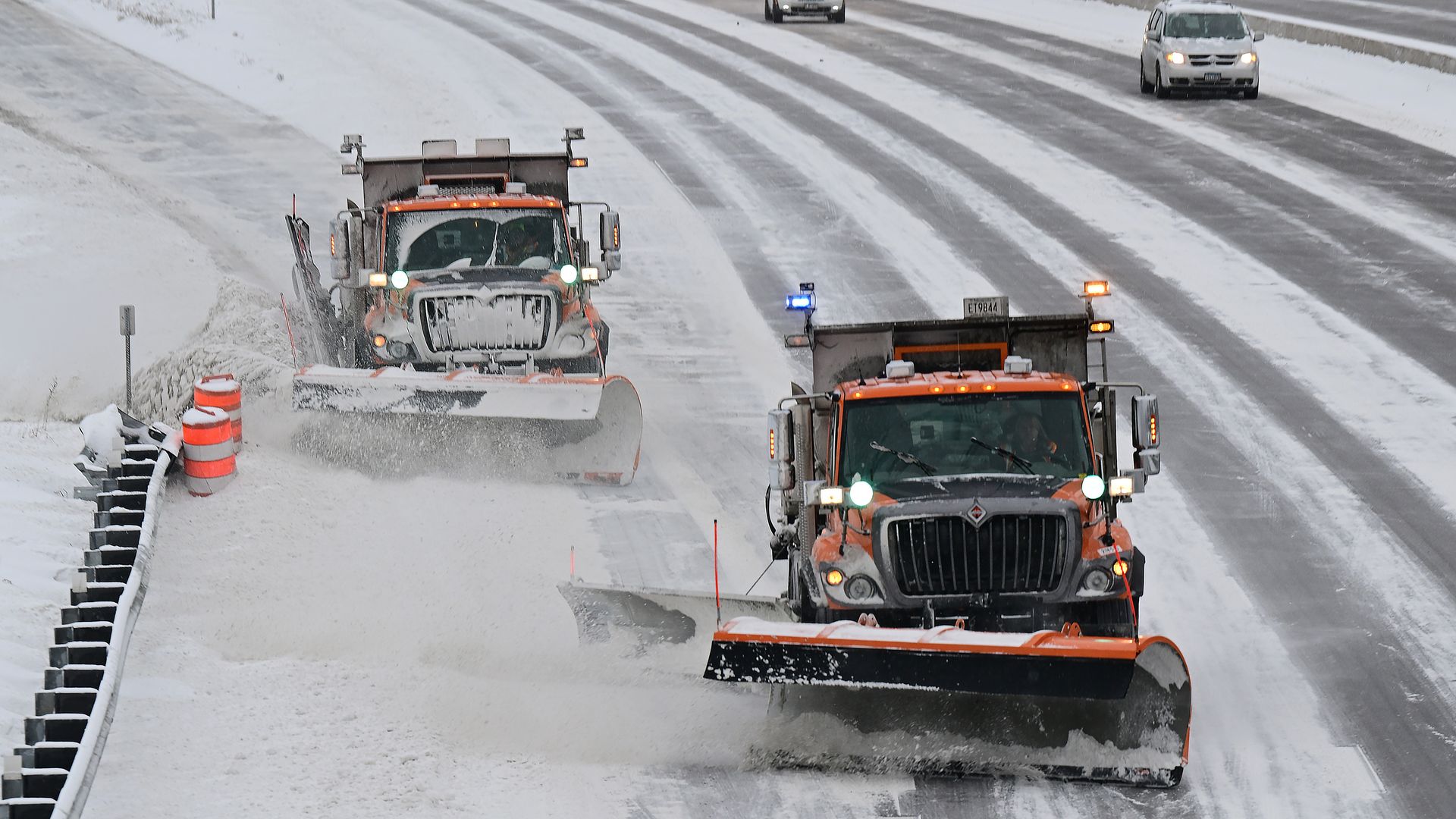 Two orange snowplow trucks clearing snow on a multi-lane highway during a snowy day with some passenger cars driving in the background.