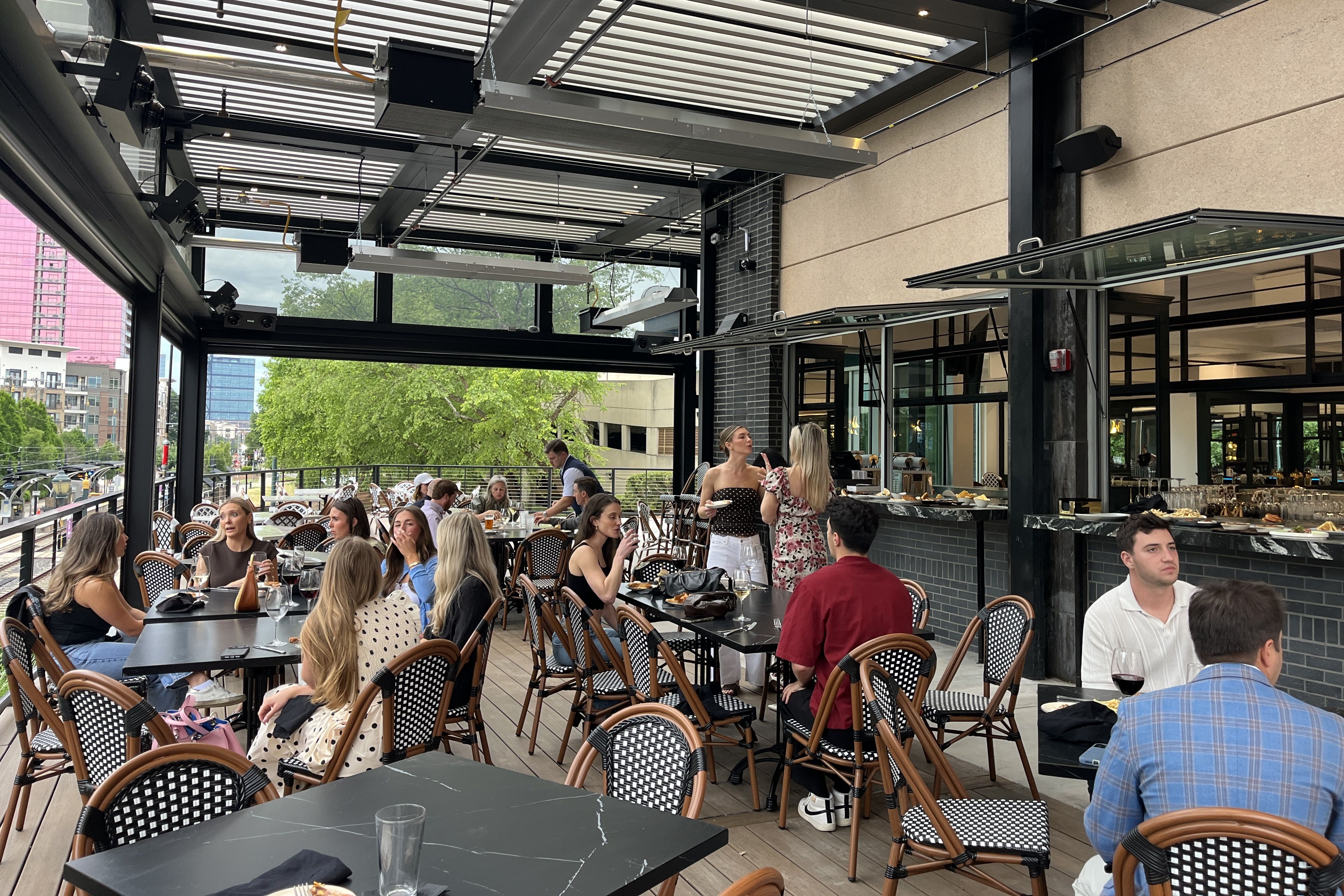 Outdoor restaurant terrace crowded with diners at black marble tables and wicker chairs. Waitstaff mingle near a serving counter under a modern industrial ceiling; glass walls reveal trees and distant buildings.