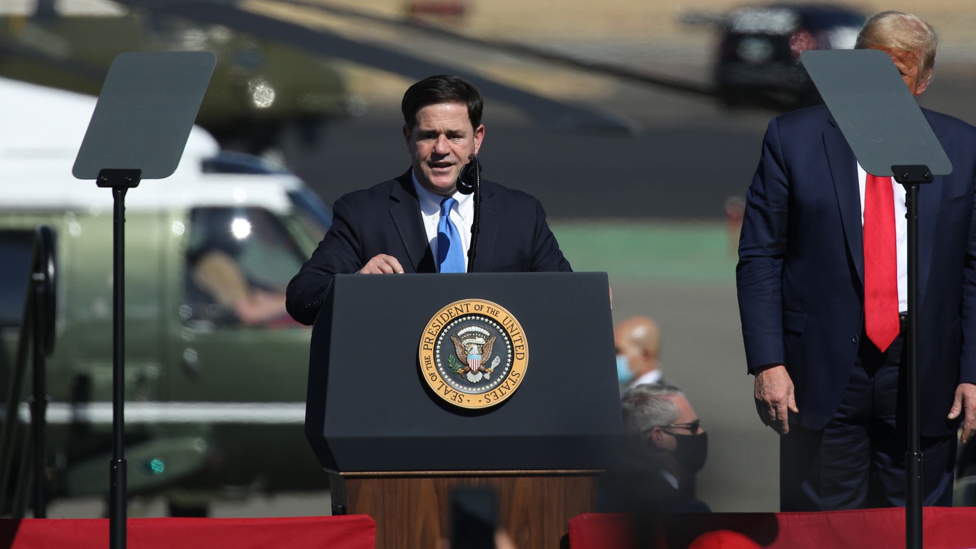A politician in a navy suit and blue tie speaks at a podium bearing the Presidential Seal outdoors, microphone in front; a second suited man stands to the right, with gray teleprompter screens nearby.