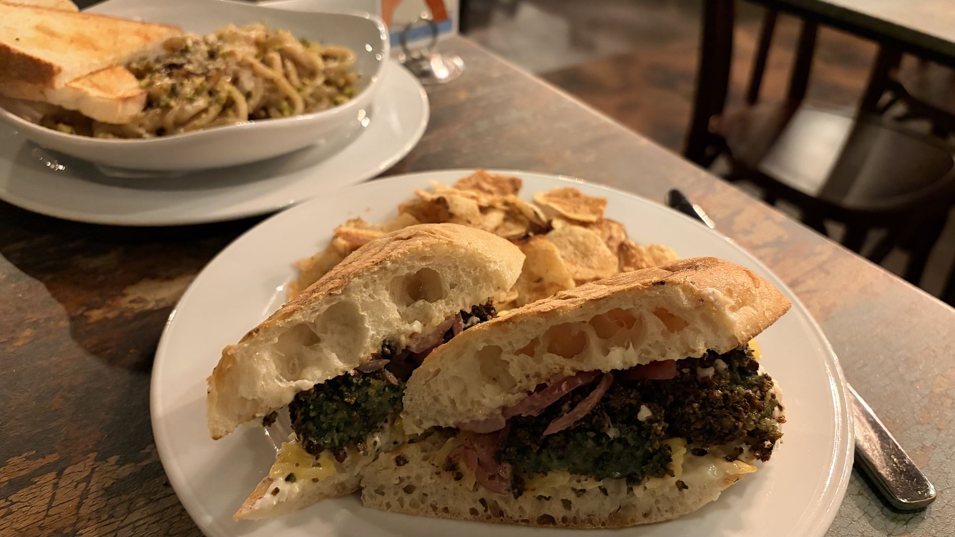 Plate with a half-cut crusty sandwich filled with greens and cheese sits on a table; a second plate in the background contains pasta with toasted bread slices.