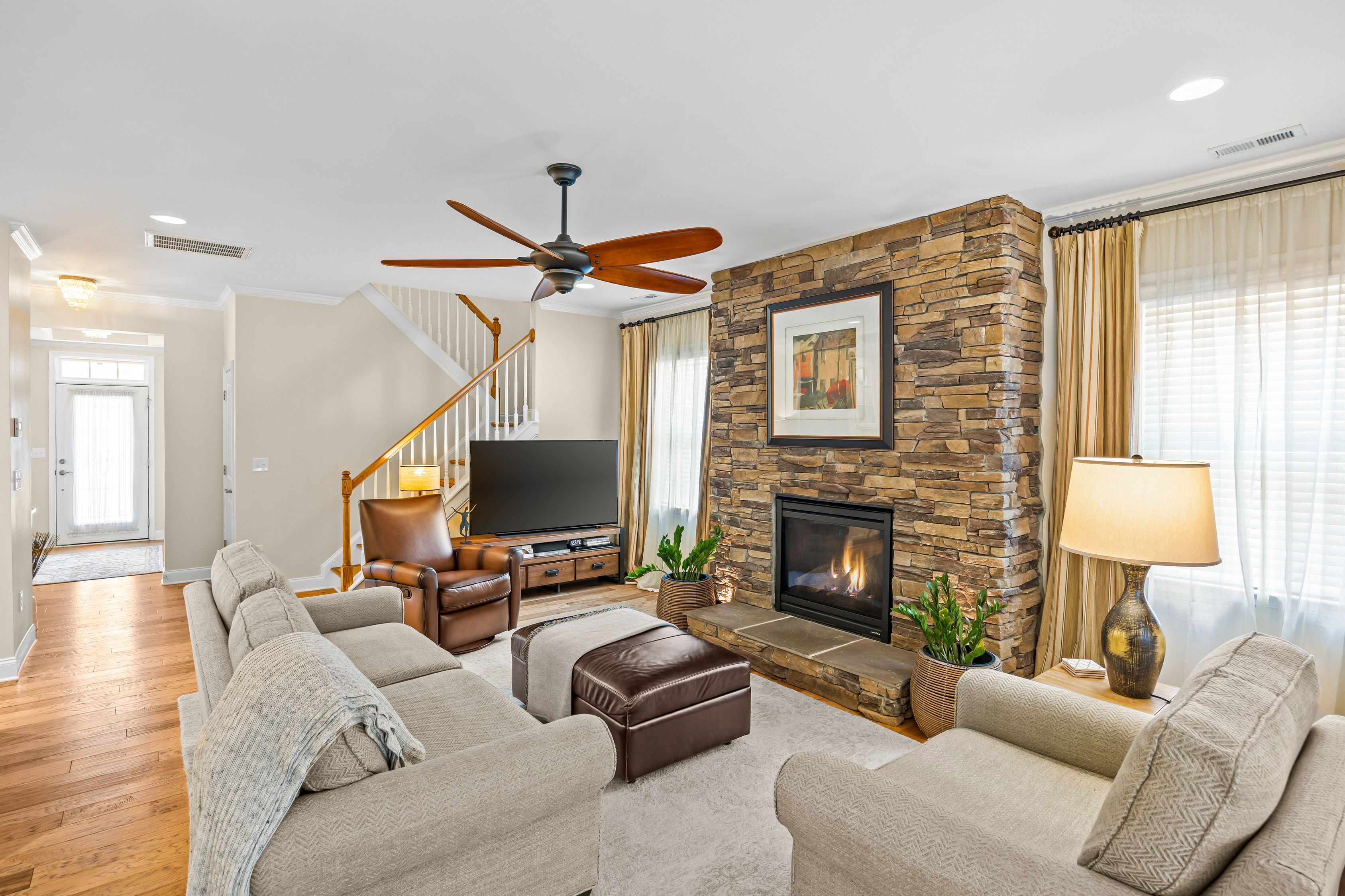 Living room featuring a stacked stone fireplace, beige walls, and a wooden ceiling fan. A gray sofa and brown leather chair face a TV on a wooden stand; large windows with beige curtains brighten the space.