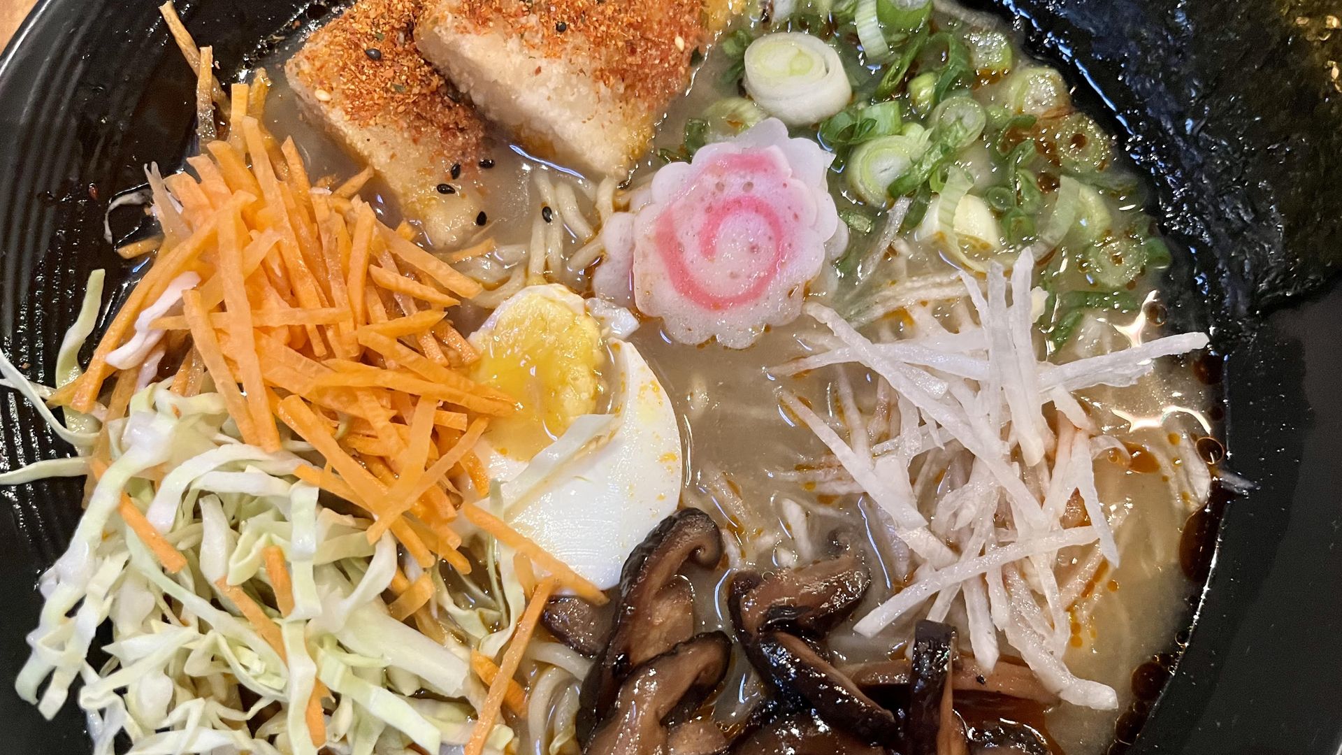 A giant bowl of ramen with fried tofu, green onions, mushrooms, cabbage, carrots, an egg, fish cakes and more.