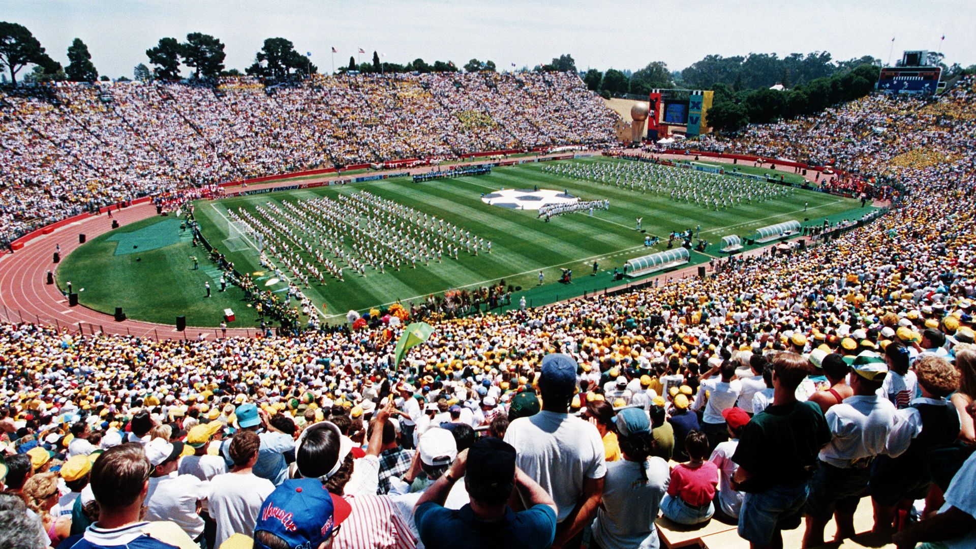A packed stadium with spectators watching a large event on the green field featuring coordinated performers and a giant star symbol, under clear blue skies.