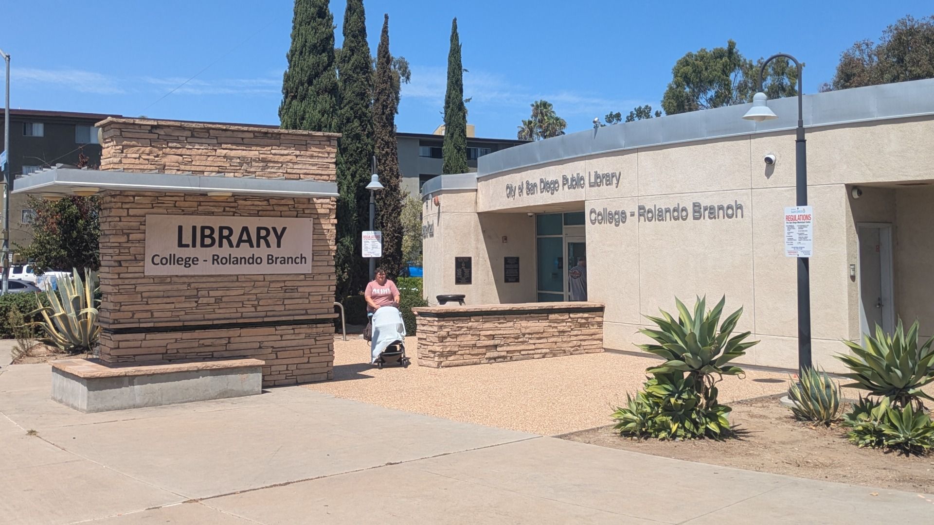 Exterior view of the City of San Diego Public Library, Rolando Branch. A stone sign reads LIBRARY College - Rolando Branch; a person pushes a stroller near the entrance on a sunny day.