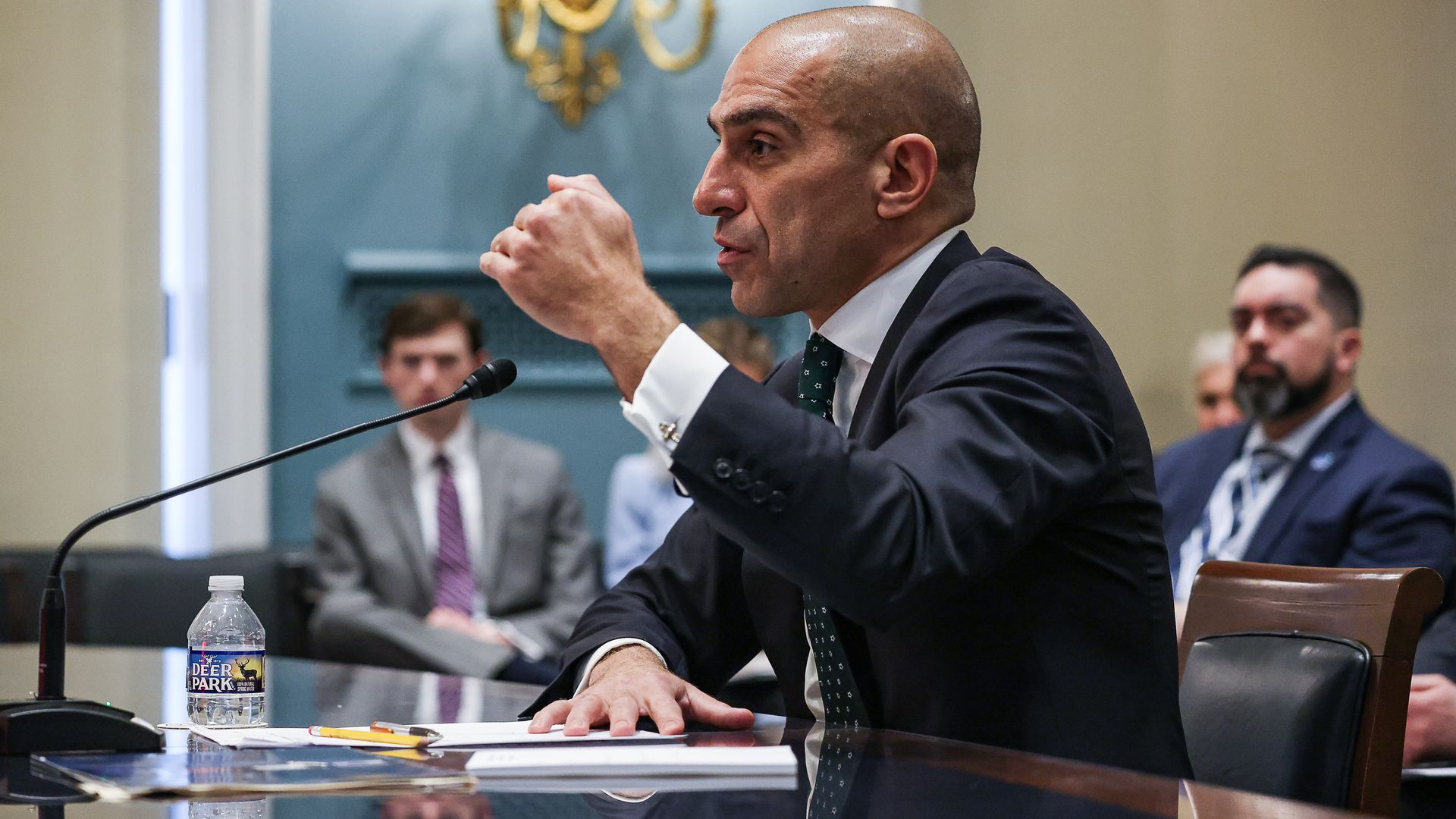 A man speaking in an official setting, with a few people blurry in the background. 