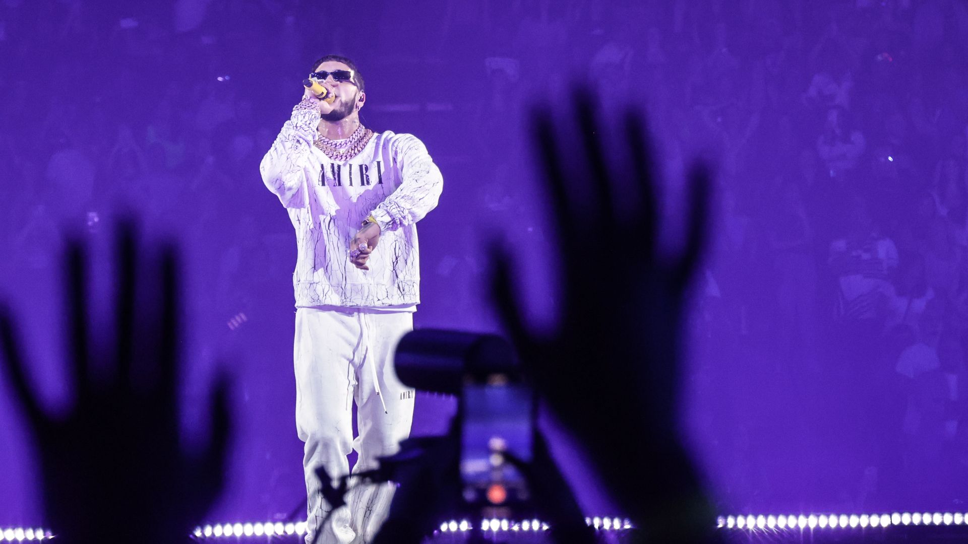 Anuel AA wears a white tracksuit and sunglasses as he sings