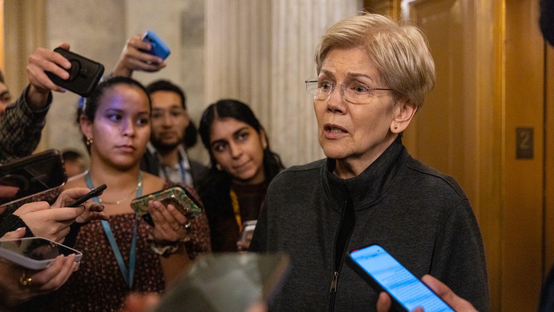 Reporters with phones surround a bespectacled short, gray-fair haired Sen. Elizabeth Warren, who's wearing a dark gray, zipup sports jacket and a black top, as she speaks.