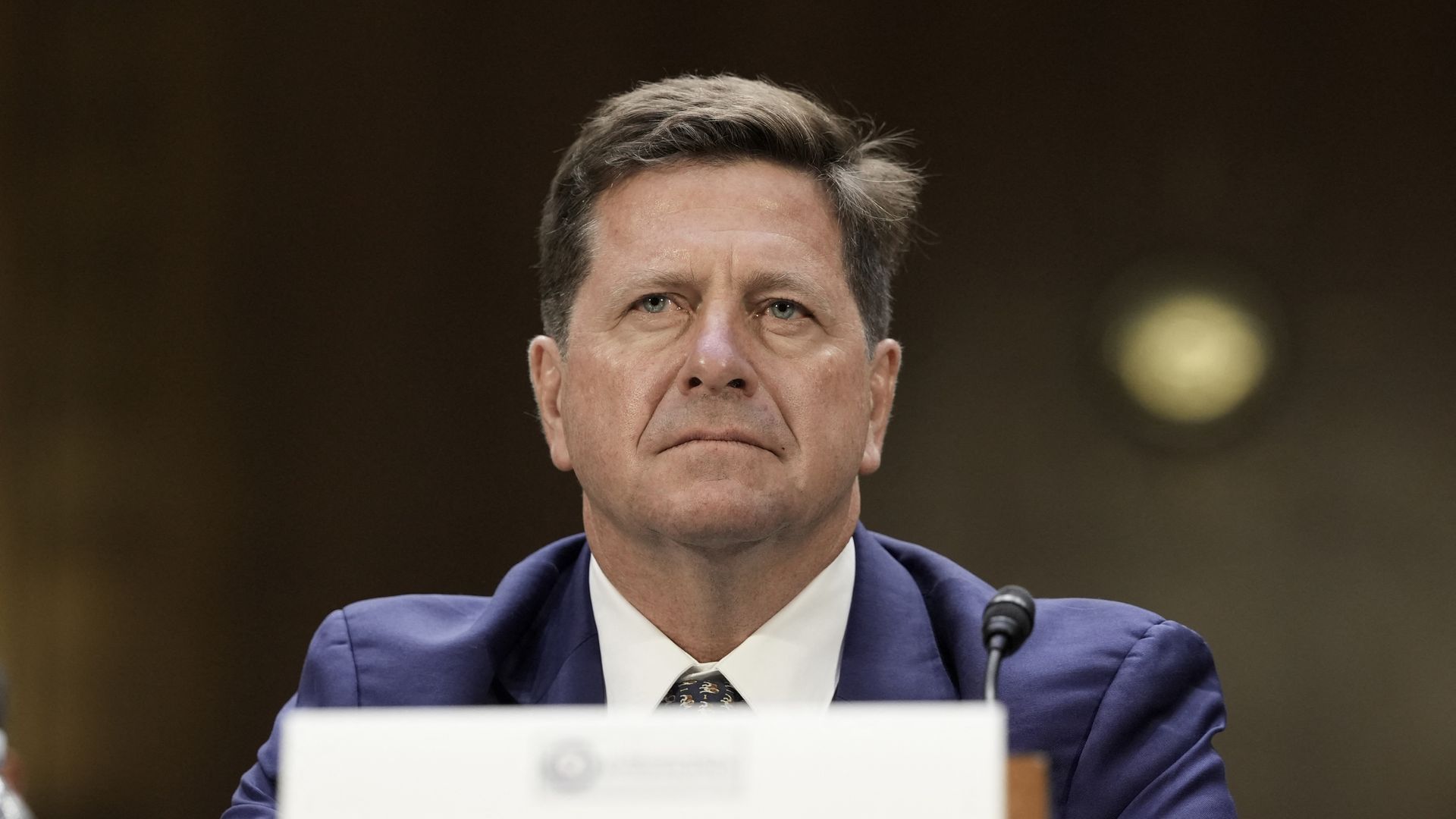 A man with brown hair who is wearing a blue suit jacket, a white collared shirt and a tie, looks on while sitting at a desk during a hearing on Capitol Hill.