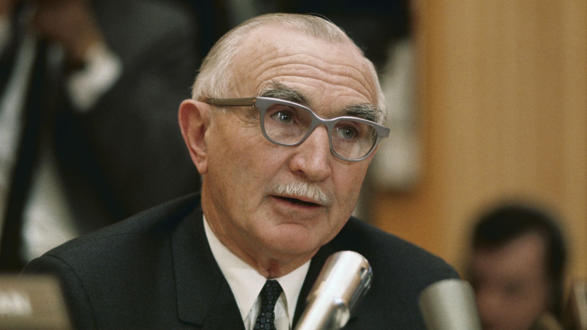 An older man with gray hair and thick eyebrows speaks into a microphone at a formal event. He wears gray-rimmed glasses, a dark suit, white shirt, and tie. The background shows other people blurred.